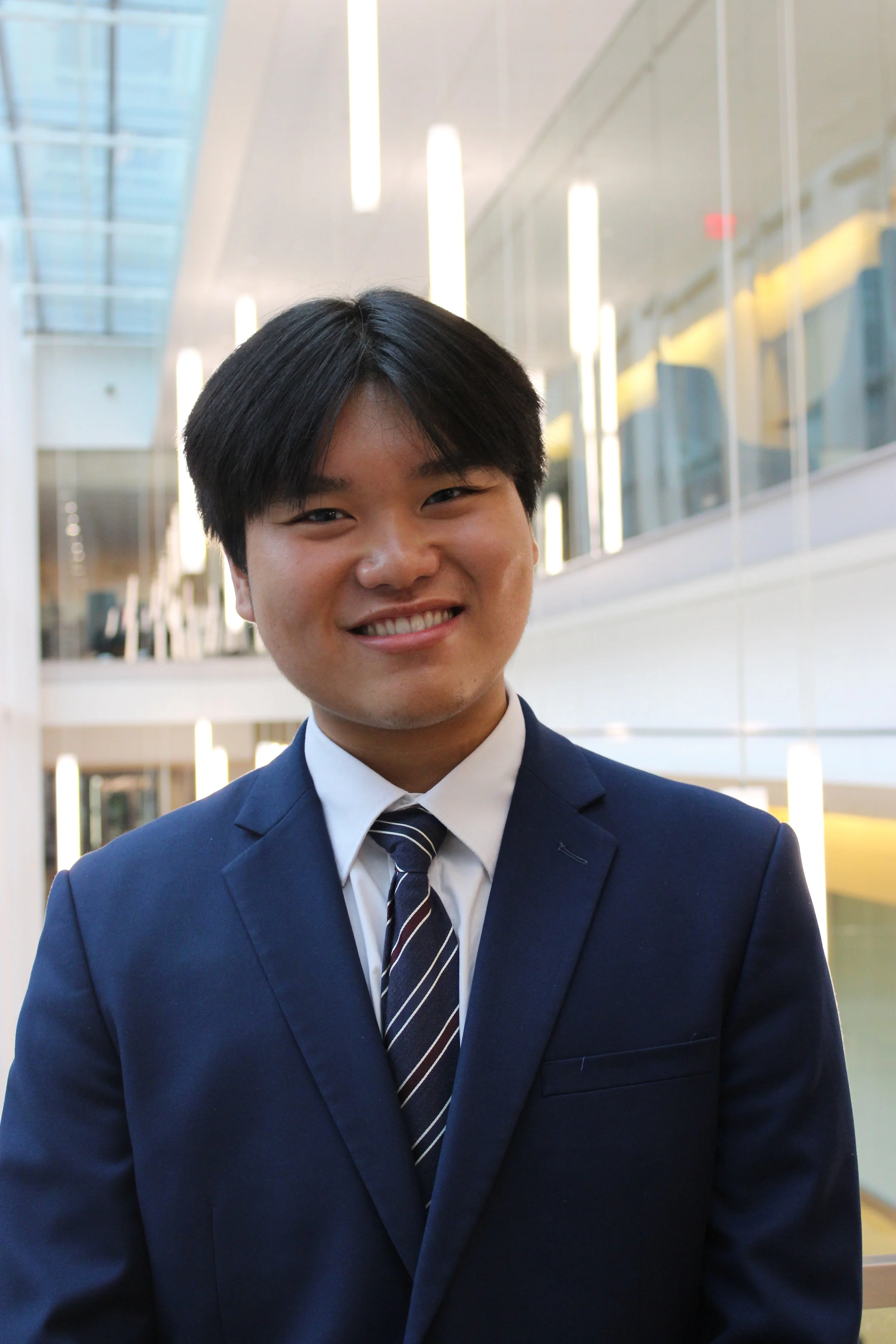 A young man in a dark blue suit, white shirt, and striped tie smiling in a modern, well-lit office building.