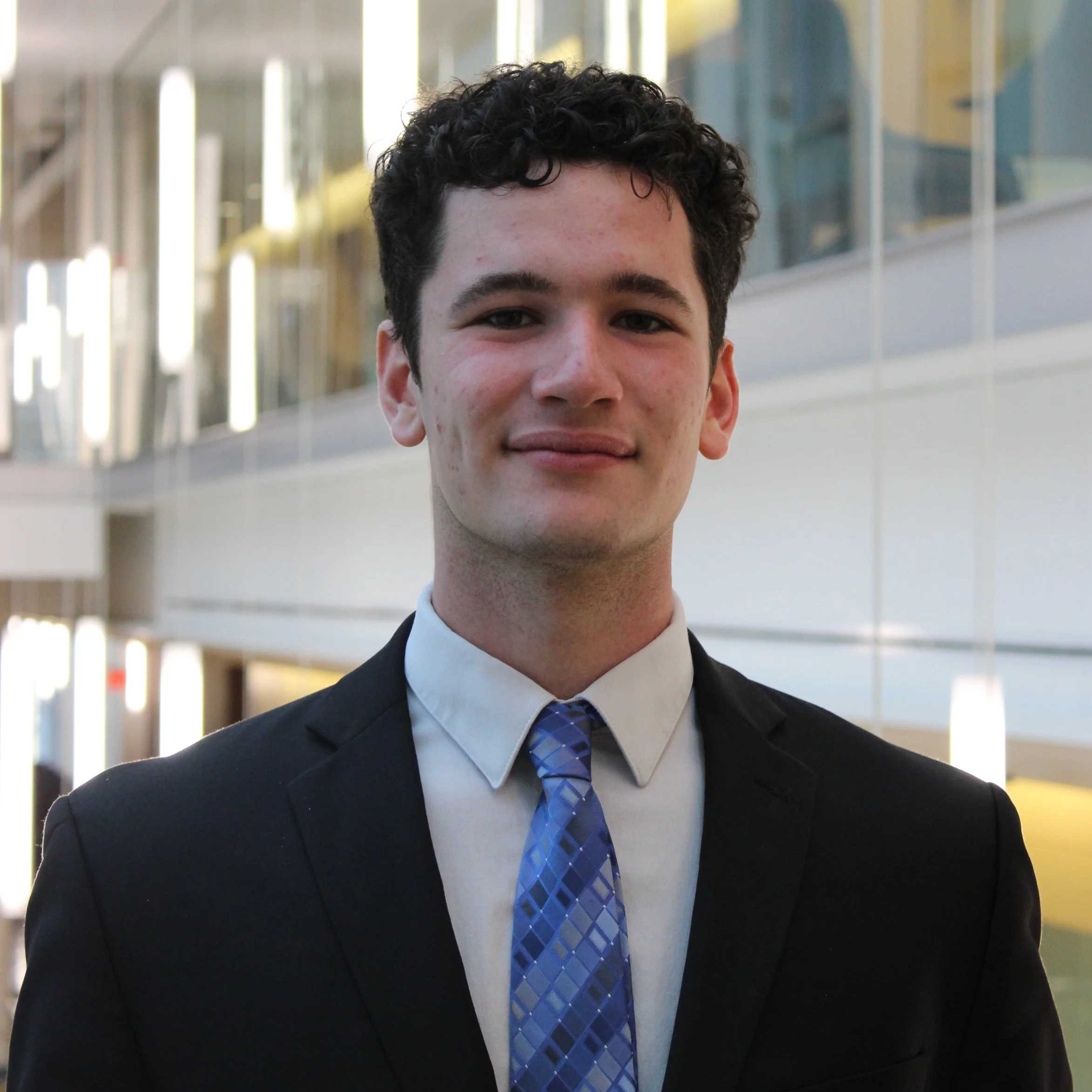 Young man in formal attire, wearing a black suit, white dress shirt, and blue patterned tie, standing indoors with a modern office or conference center background.