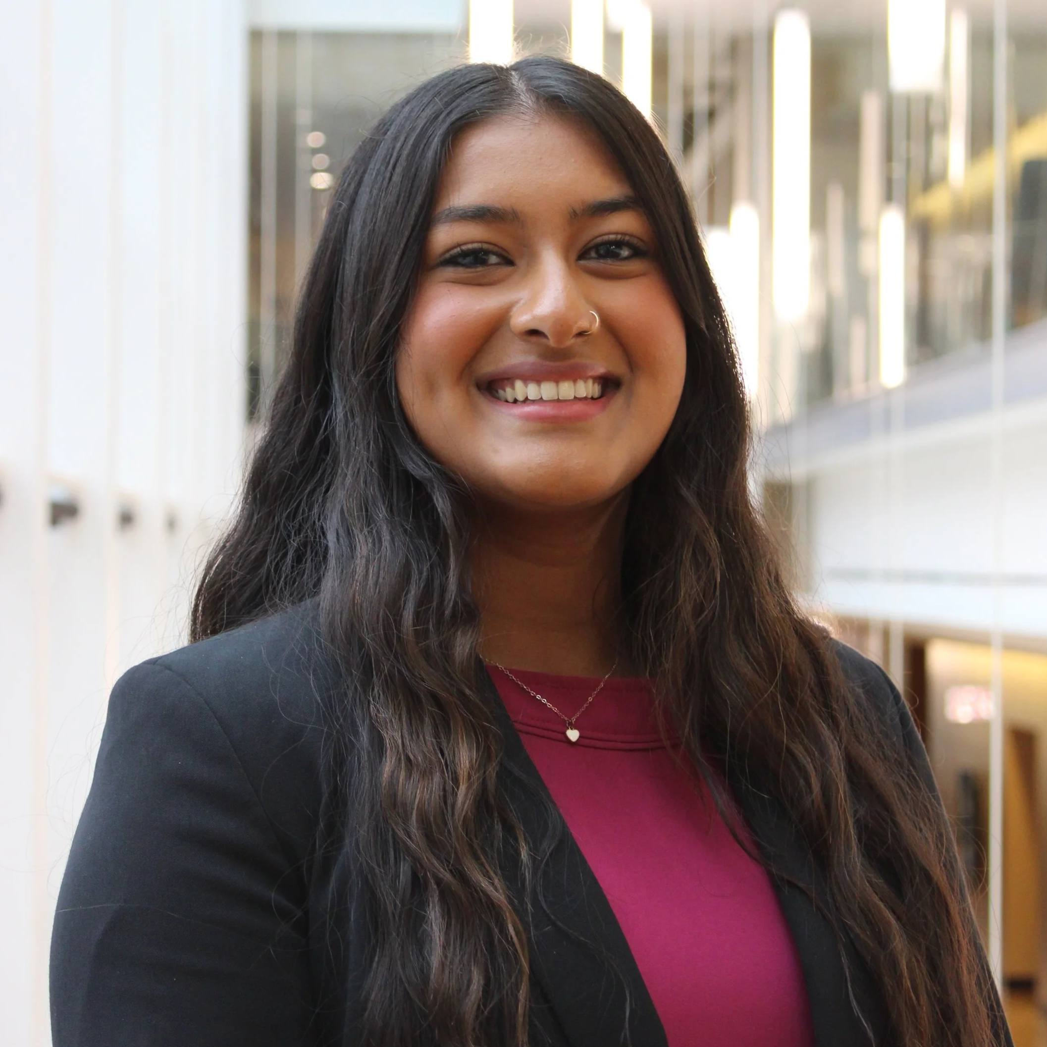 Portrait of a smiling woman with long dark hair, wearing a black blazer and a red top, standing in an indoor location with mirrored walls.