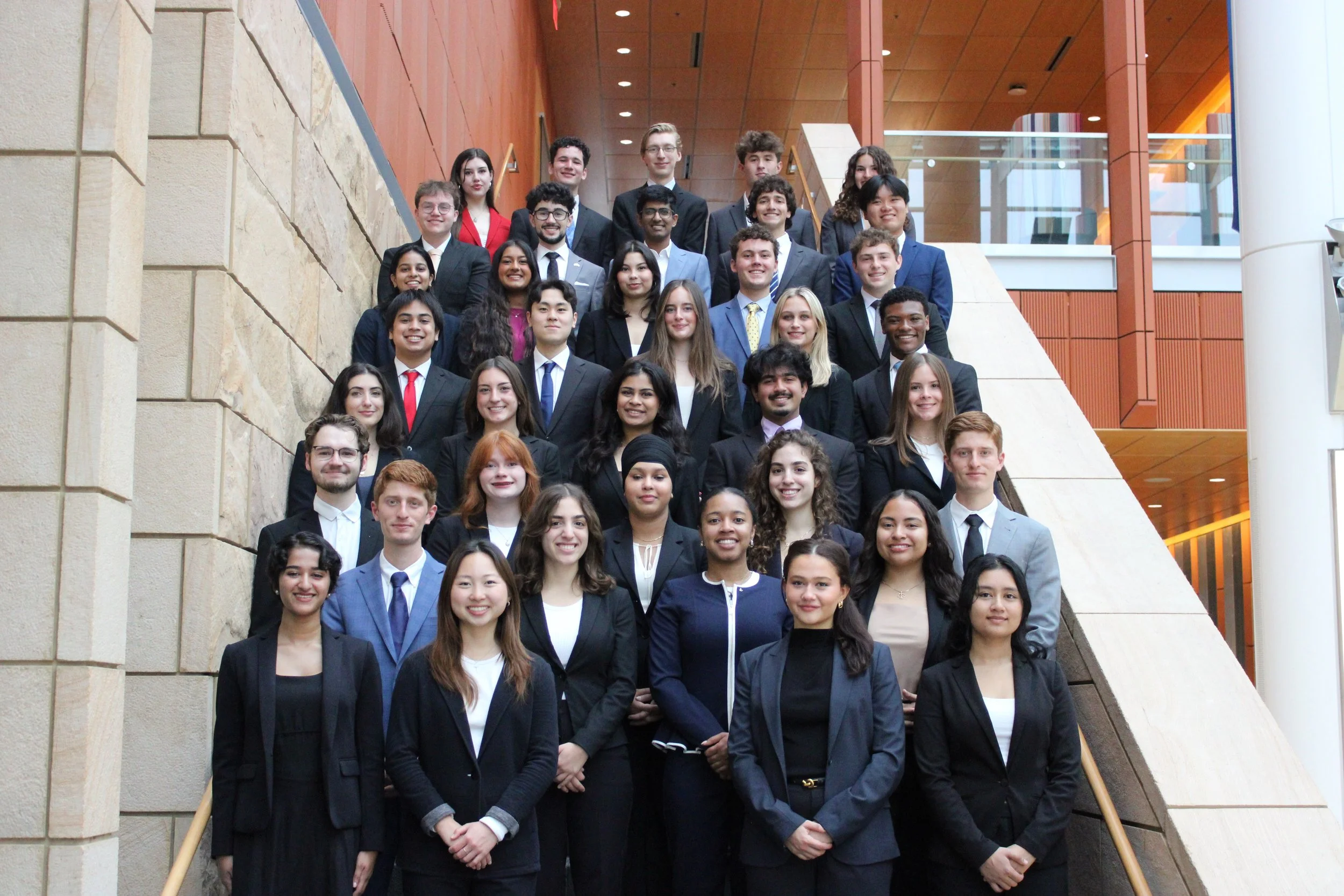 Group of young adults in business attire posing on a staircase inside a modern building.