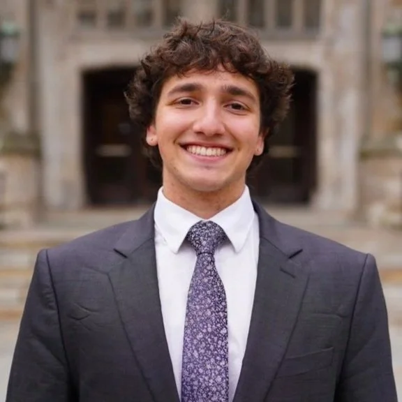Young man in a dark suit and tie smiling outdoors in front of a stone building.