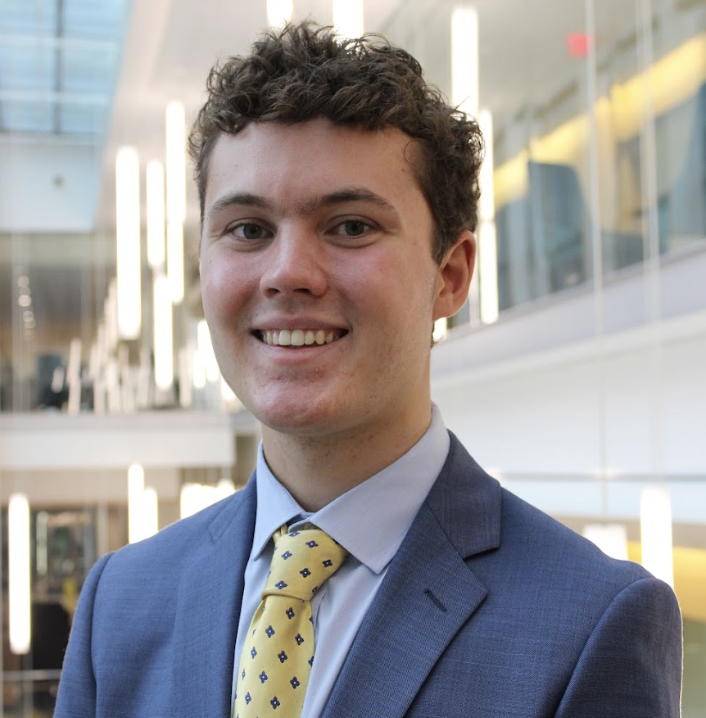 A young man with curly brown hair and fair skin is smiling at the camera. He is wearing a blue suit, a light blue shirt, and a yellow tie with small black patterns. The background shows a modern building with reflective glass windows and interior lighting.