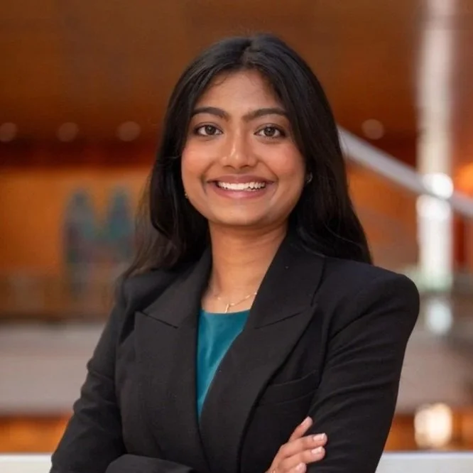 Professional woman with long black hair smiling, wearing a black blazer and teal top, standing indoors with a blurred background.