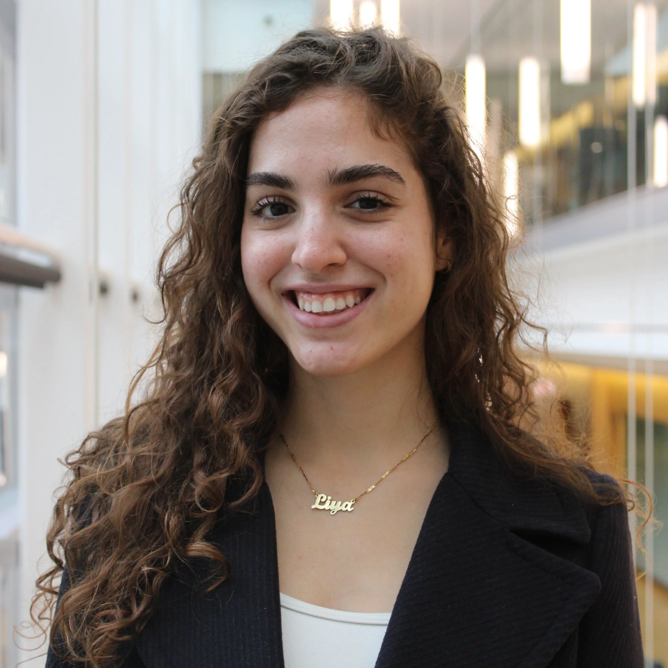 A young woman with curly brown hair, smiling, wearing a black blazer, white top, and a gold necklace with the name 'Liya' written on it, standing indoors in a modern building with glass and white walls.