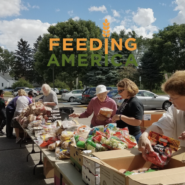 People volunteering at a food distribution event with tables filled with bread and groceries, outdoors on a sunny day.