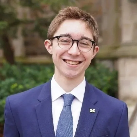 Young man in glasses smiling, dressed in a blue suit and tie, outdoors with greenery in the background.