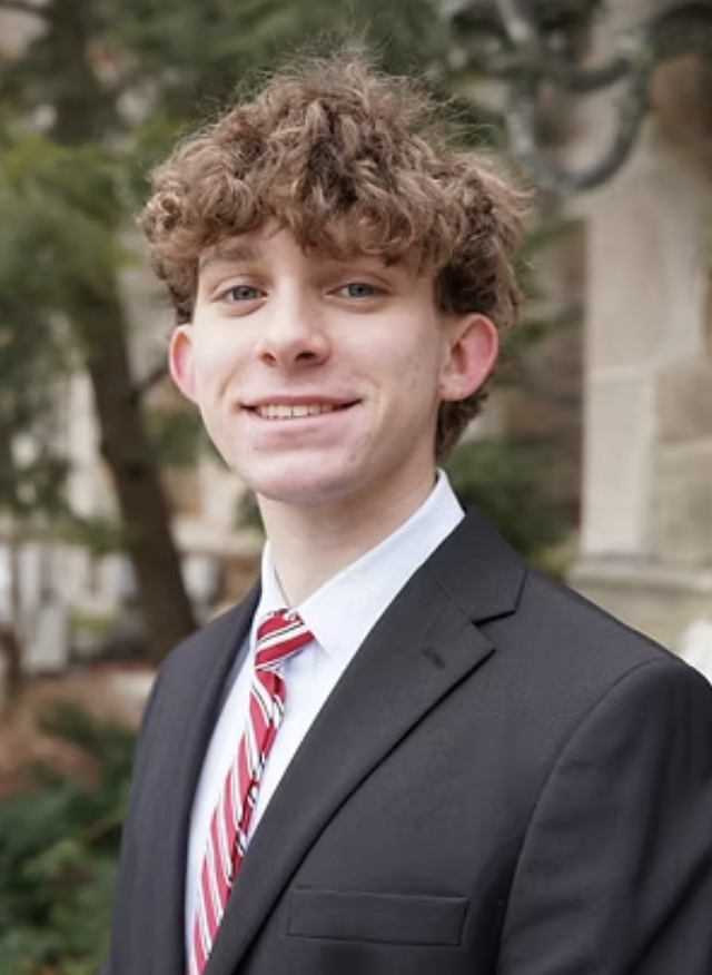 Young man with curly brown hair wearing a black suit, white shirt, and red-striped tie, smiling outdoors with trees and a building in the background.