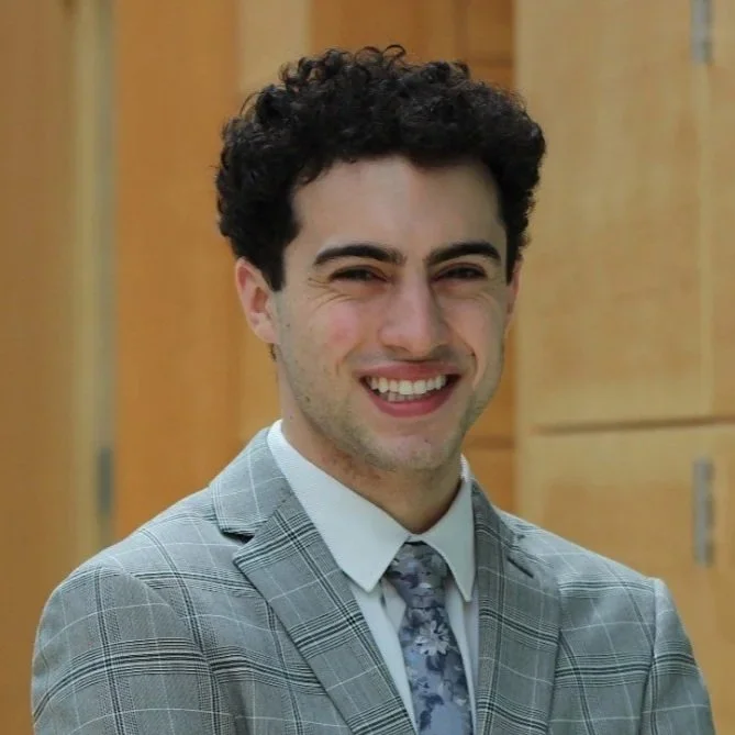 A young man with curly dark hair, smiling, wearing a gray plaid suit, white shirt, and gray patterned tie, standing in front of wooden lockers.