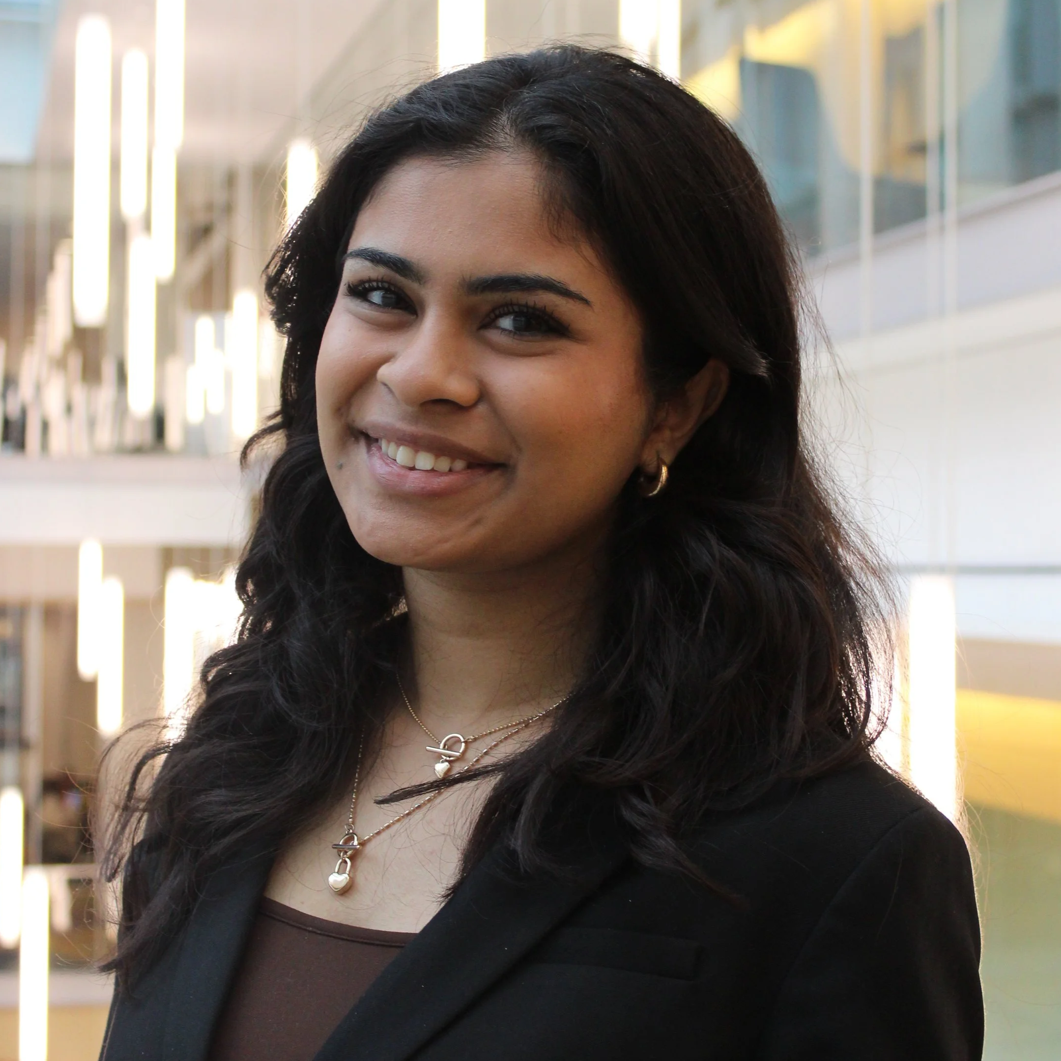 A young woman with dark wavy hair smiling at the camera in a modern indoor setting with glass and metal architecture.