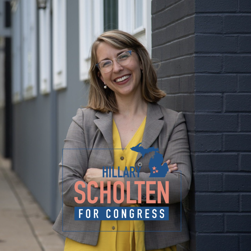 A woman with glasses and shoulder-length hair standing outdoors, smiling, with her arms crossed, next to a dark brick wall. She is wearing a grey blazer over a yellow top. The image features a campaign sign for Hillary Scholten for Congress, with a graphic of the state of Michigan.