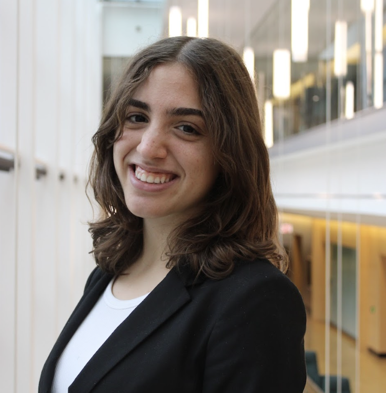 Smiling young woman with shoulder-length brown hair, wearing a black blazer and white shirt, standing indoors in a modern building with glass walls and warm lighting.