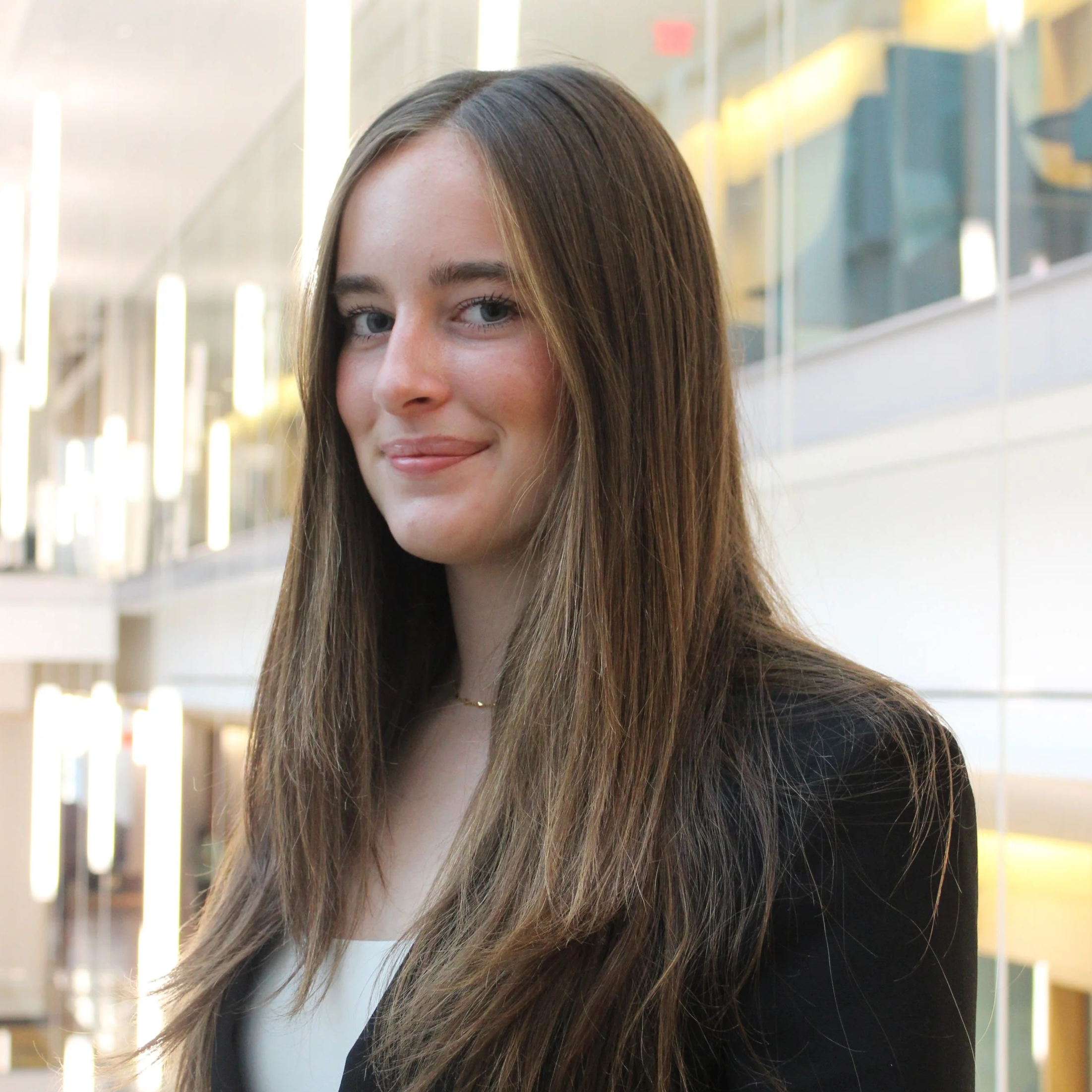 A young woman with long brown hair and a slight smile, wearing a black blazer and a white top, standing in an indoor modern office building with glass walls and bright lighting.