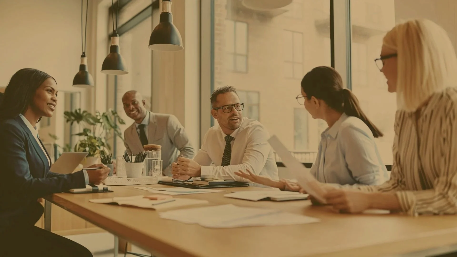 A diverse group of five people engaged in a discussion during a meeting in a modern office with large windows and hanging pendant lights.