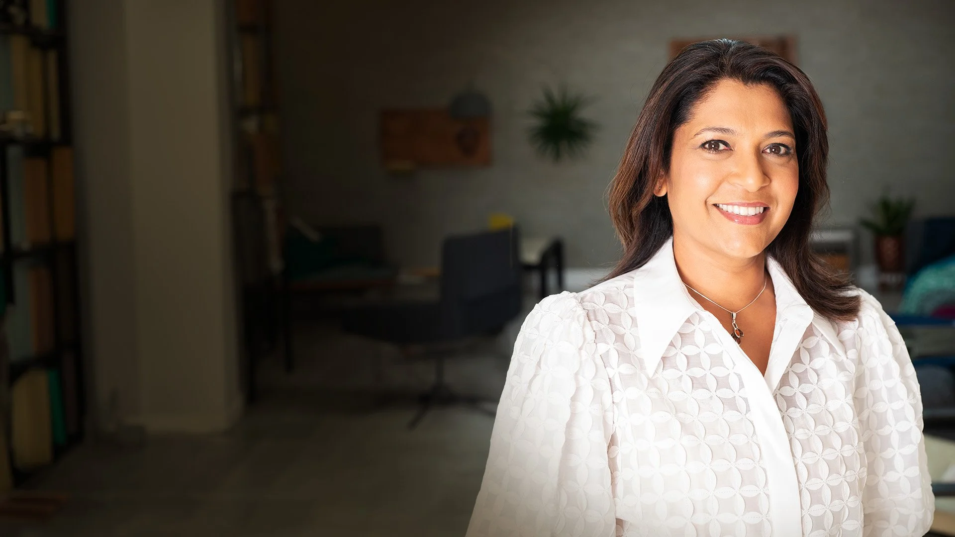 Navita Doshi with brown hair and a white patterned blouse, smiling indoors, with a blurred background of a home office or living space.