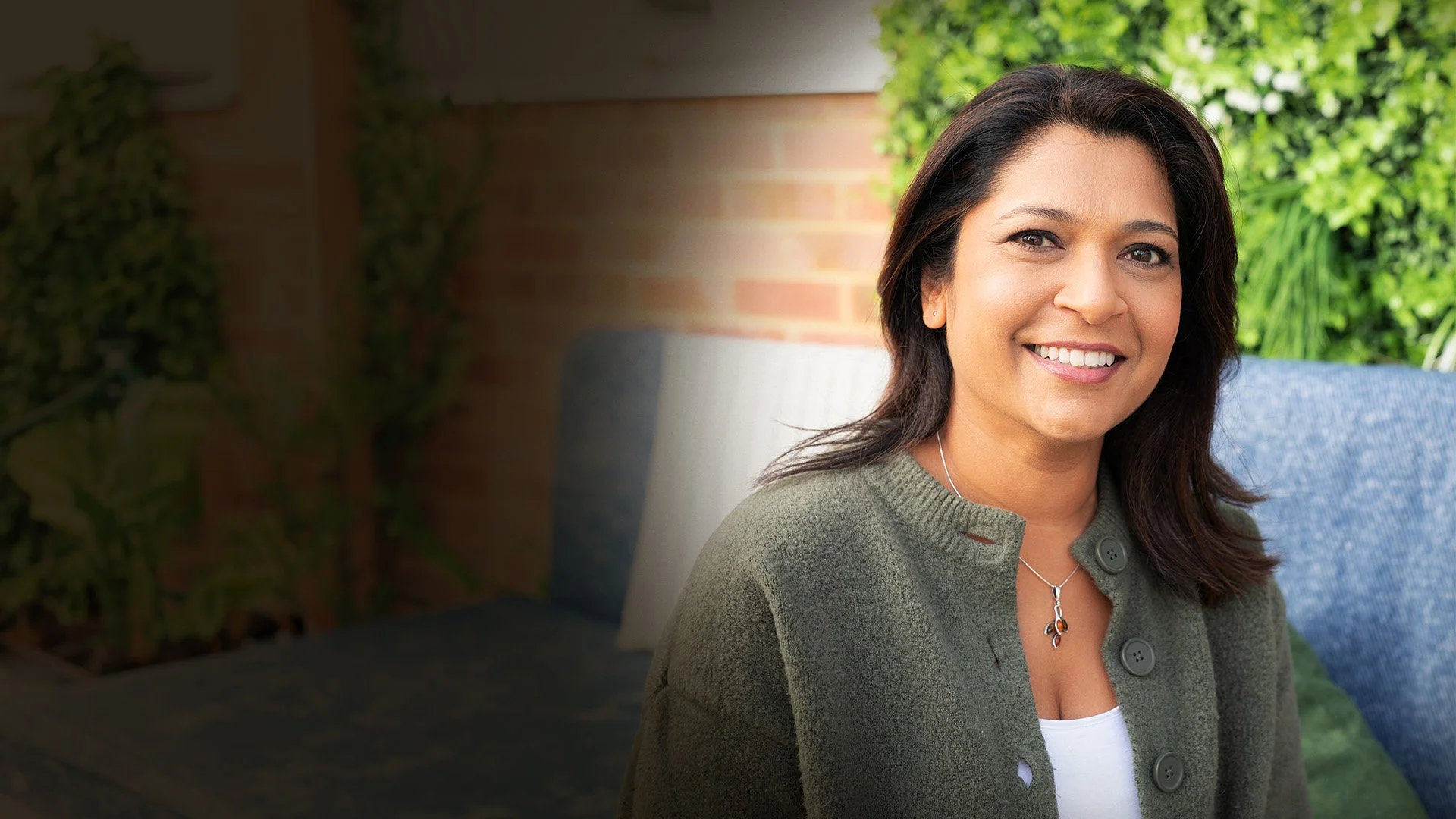 Navita Doshi with shoulder-length dark hair, smiling, wearing a green cardigan and a silver necklace, sitting outdoors with greenery and a brick wall in the background.