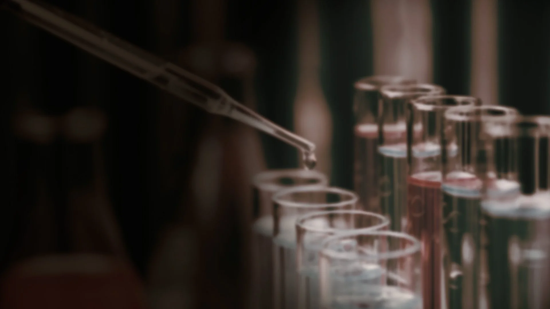 Close-up of a pipette adding a drop of liquid to test tubes in a laboratory setting.