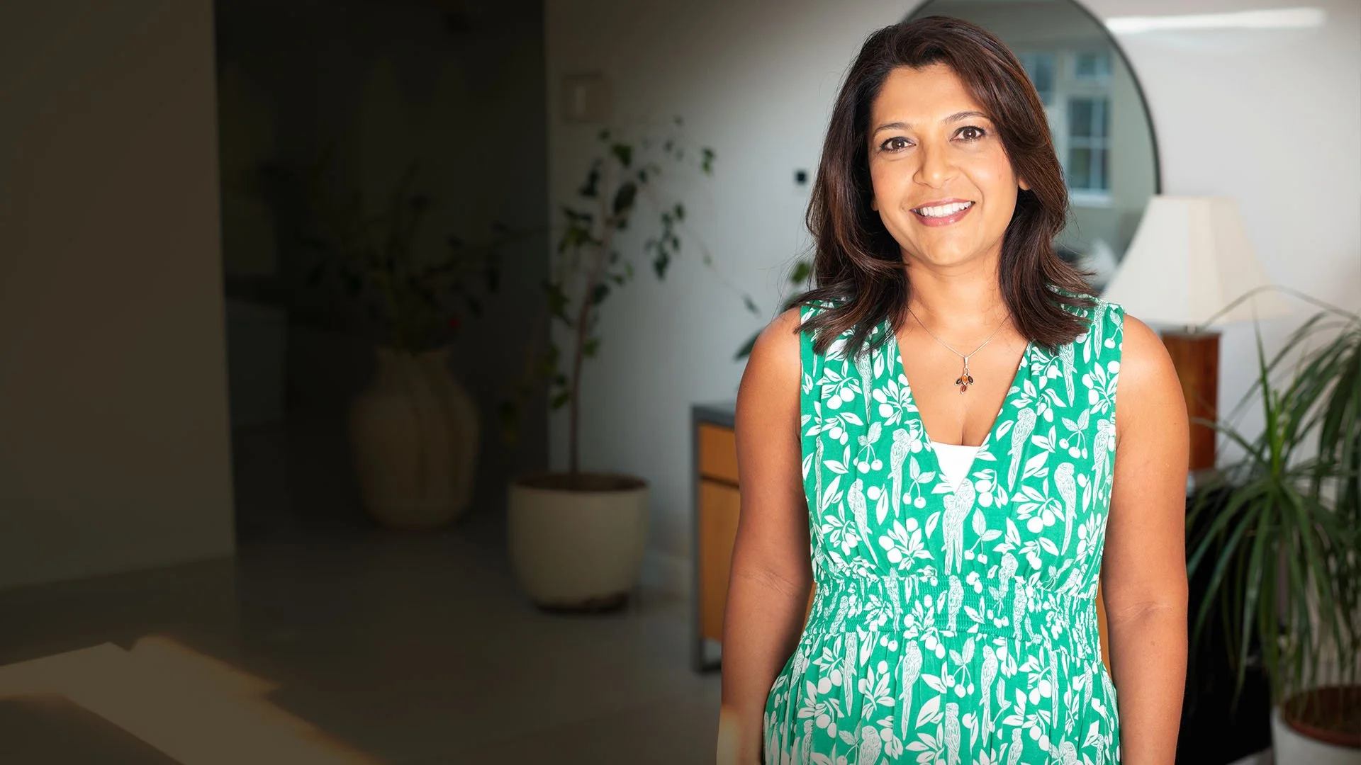Navita Doshi, with shoulder-length dark brown hair, smiling, wearing a green sleeveless dress with white bird and leaf patterns, standing indoors with a plant and a lamp in the background.