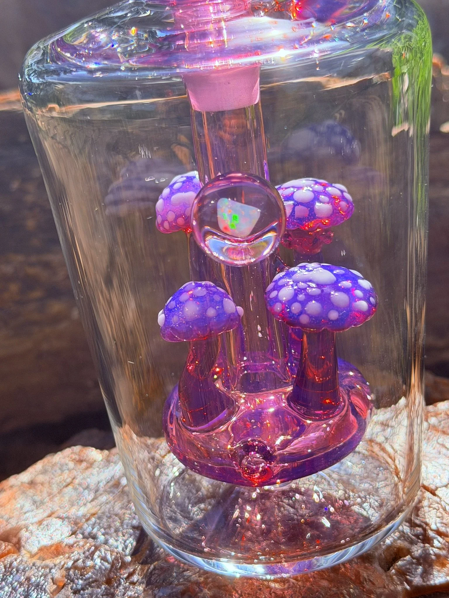 Close-up of a glass sculpture with abstract pink, purple, and green colors, featuring mushroom-like forms with white spots, placed on a textured rocky surface.