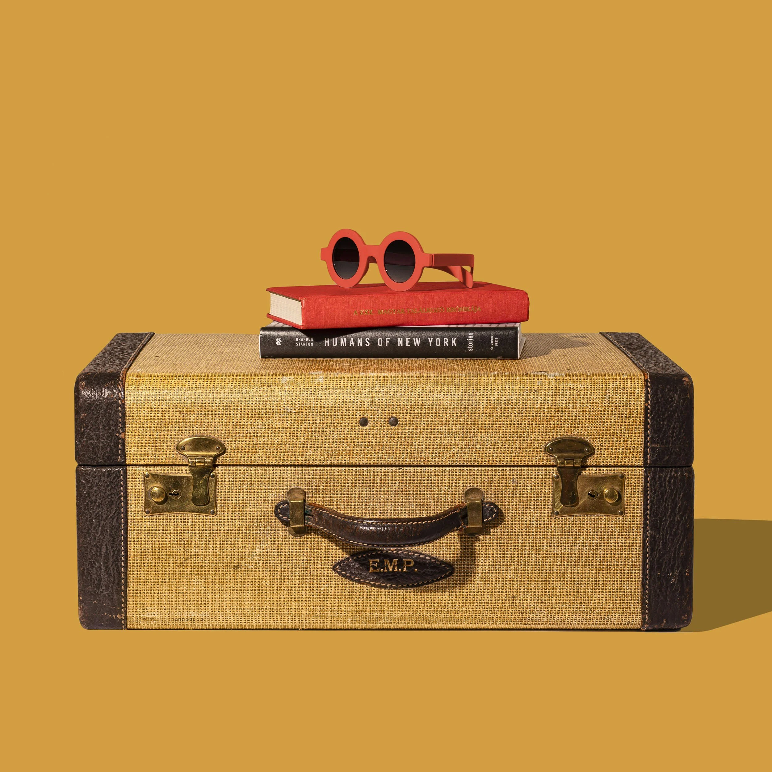 A vintage suitcase with a textured fabric surface, leather accents, and metal locks, with a handle and an embossed tag reading E.M.P.  on top of it against a plain brown background, with three books and red sunglasses placed on the books.