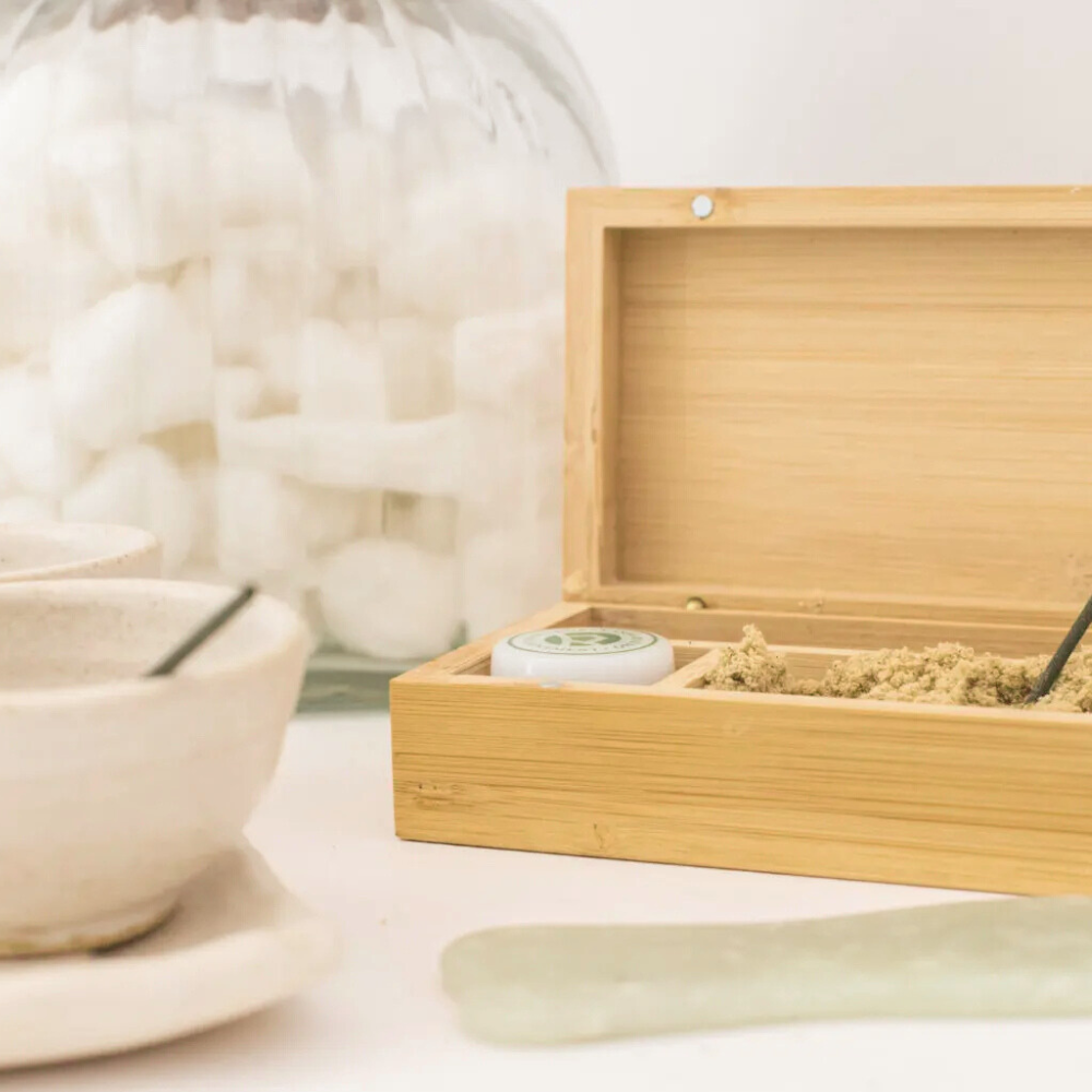 An open wooden box containing a white container and beige powder, with tea spoons, ceramic bowls, and a large glass jar in the background on a white surface.