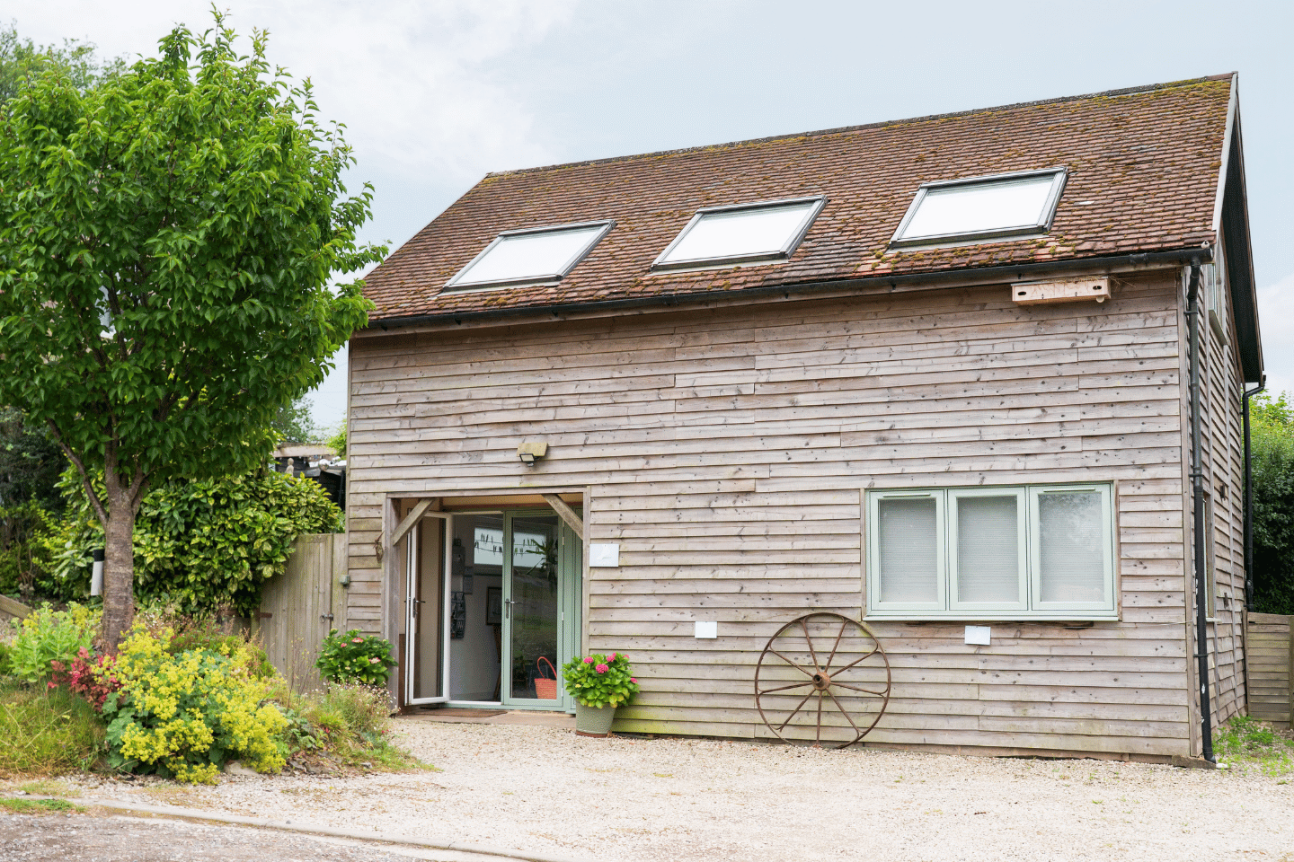 A two-story wooden house with a brown tiled roof, three skylights, and a sliding glass door at the entrance, surrounded by greenery and decorative garden items.