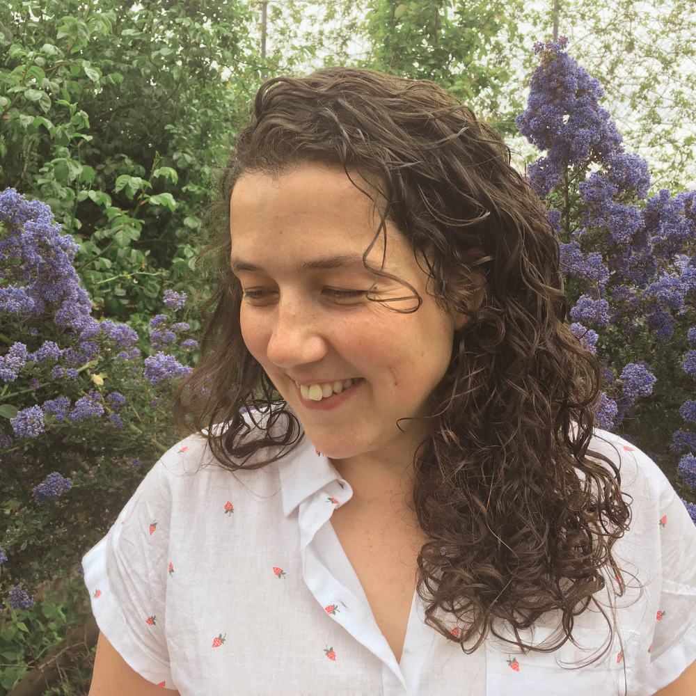 A young woman with curly brown hair smiling outdoors surrounded by purple flowers and green foliage.