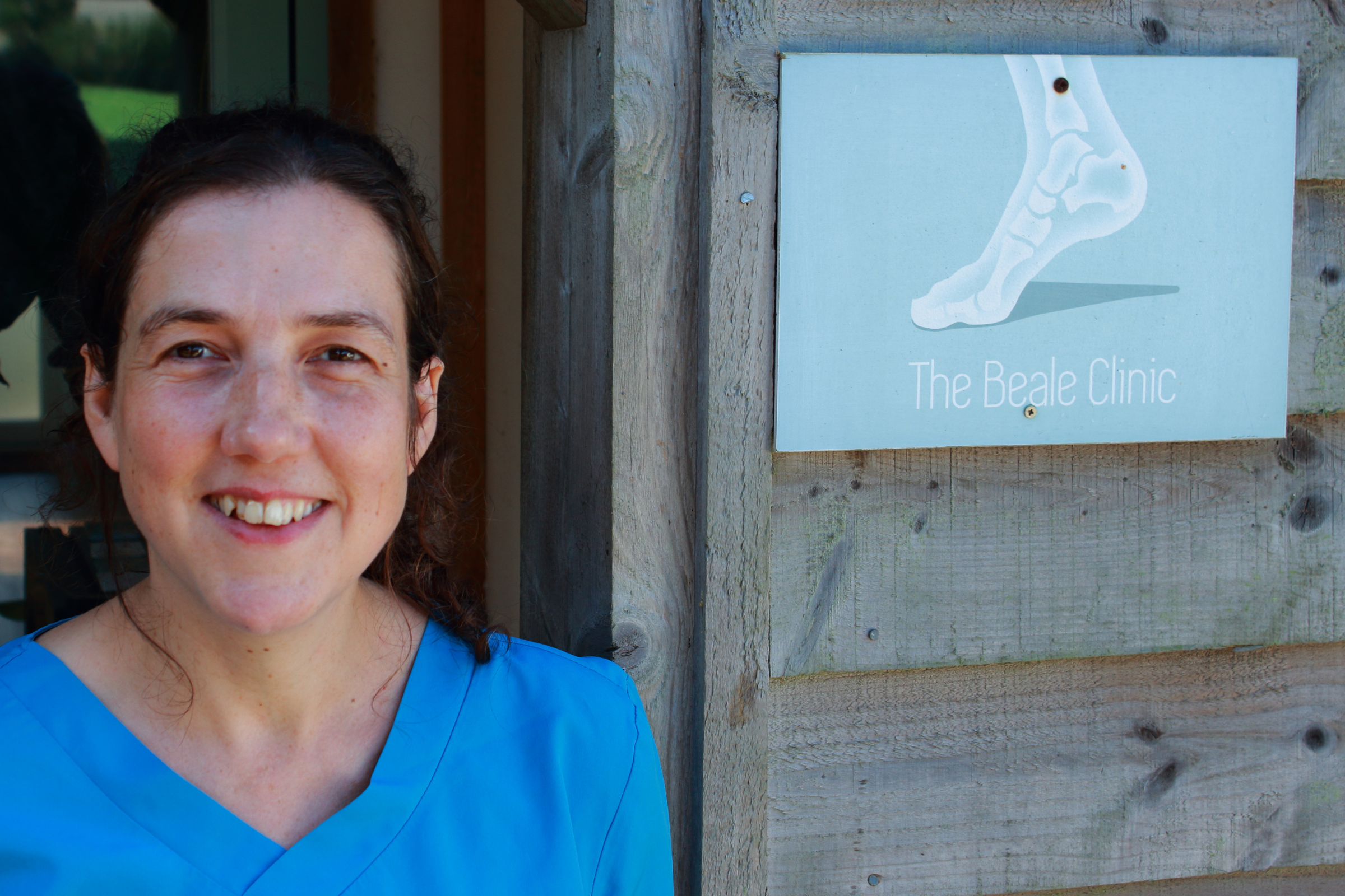 A woman (Elizabeth Nicholson, Acupuncturist) in blue scrubs standing outside The Beale Clinic, smiling at the camera, with a sign showing an illustration of a foot and the clinic's name.