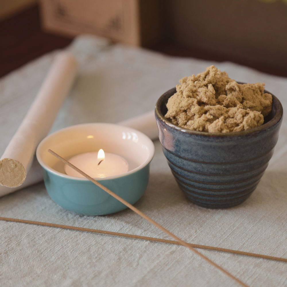 A lit candle in a small teal bowl, a large bowl filled with a beige powdered substance, a rolled paper tube, and incense sticks on a white cloth surface.