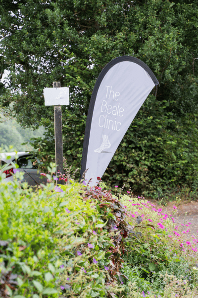 A gray feather-shaped sign reads "The Beale Clinic" and stands near green bushes and pink flowers with a large tree in the background.