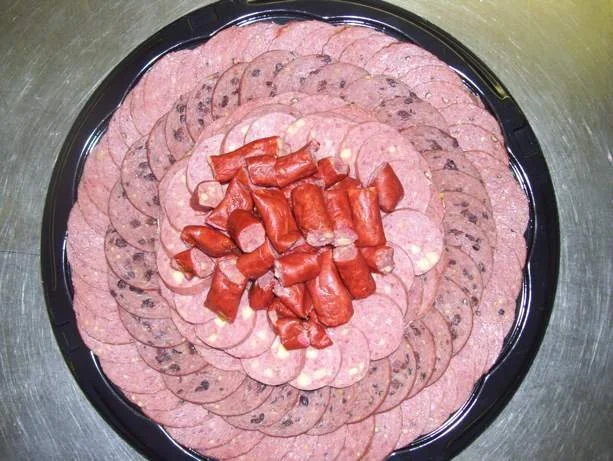 A black platter with various sliced deli meats, including pepperoni and ham, arranged in concentric circles with some chunks of cooked bacon or sausage in the center.