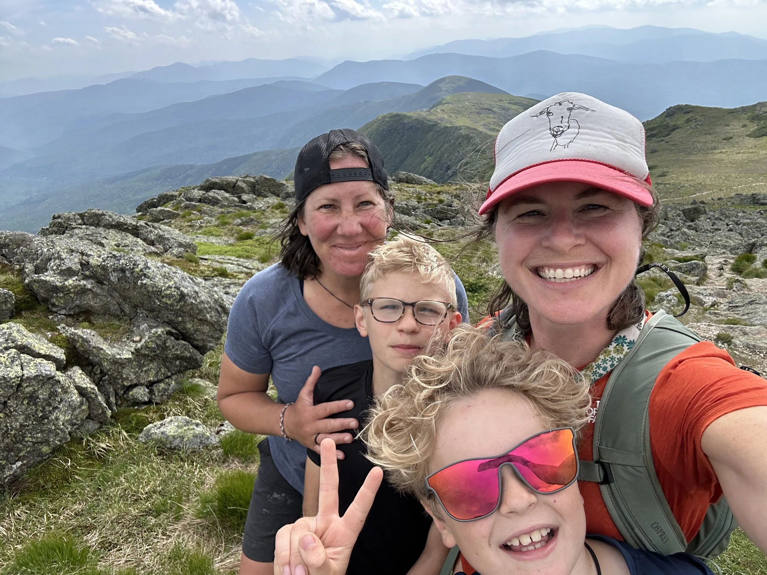 Smiling family of four taking a selfie on a mountain trail with a scenic view of rolling green mountains under a partly cloudy sky.