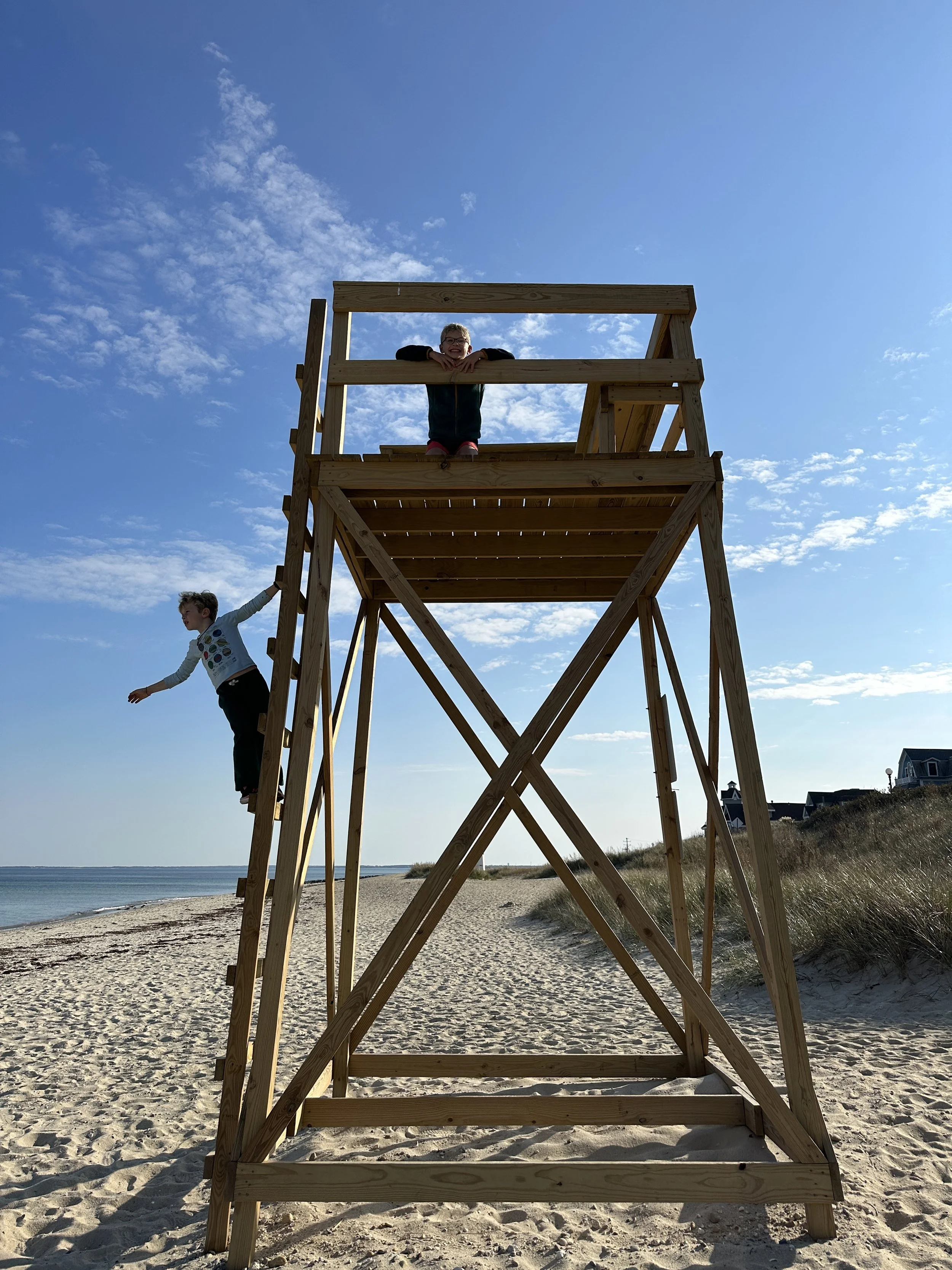A wooden lifeguard tower on a sandy beach with a boy hanging from the side, and another boy smiling from the top of the tower, under a blue sky with scattered clouds.