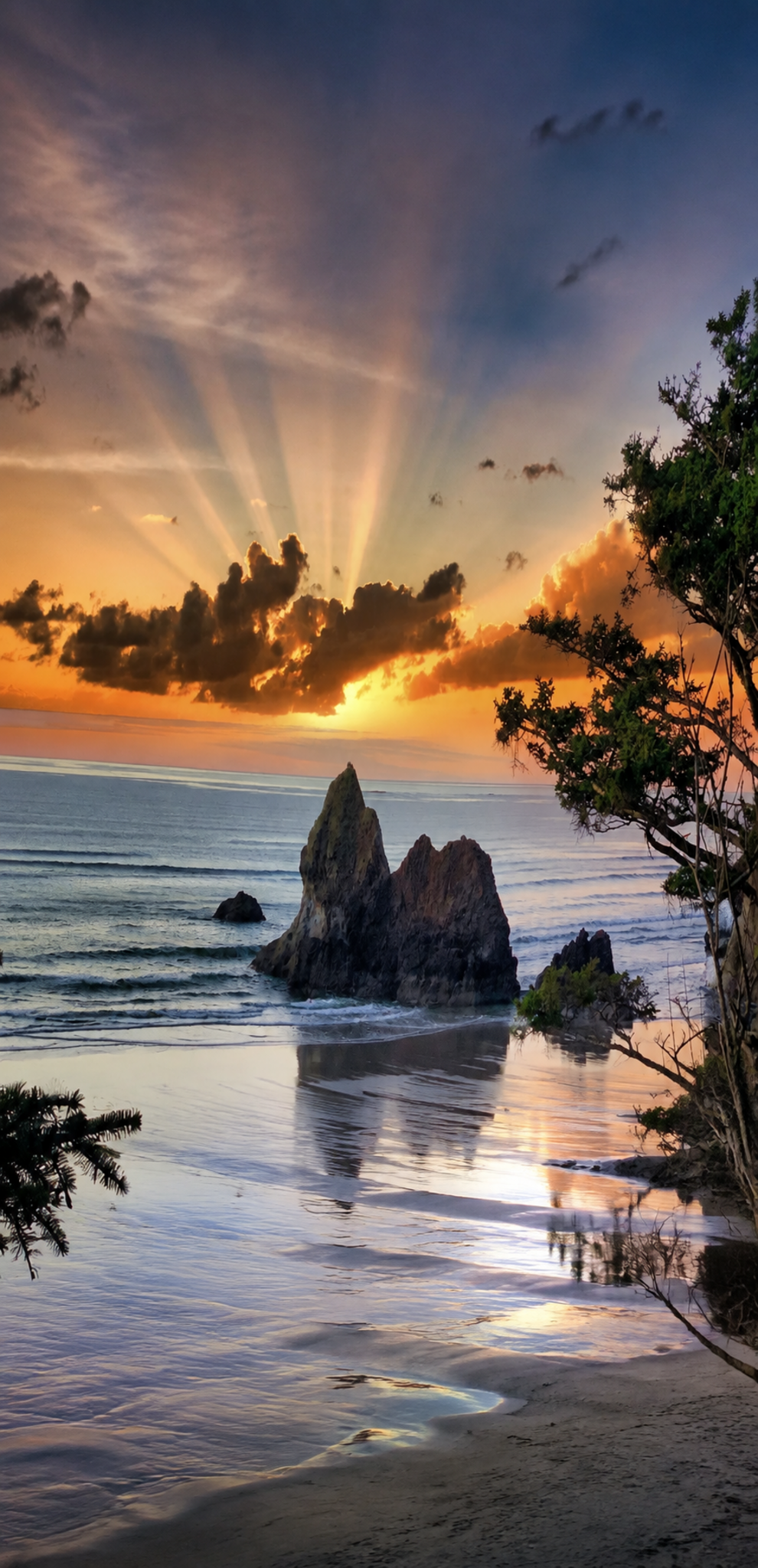 Sunset over the ocean with rays of sunlight breaking through the clouds, large rocks in the water, and trees along the shoreline.