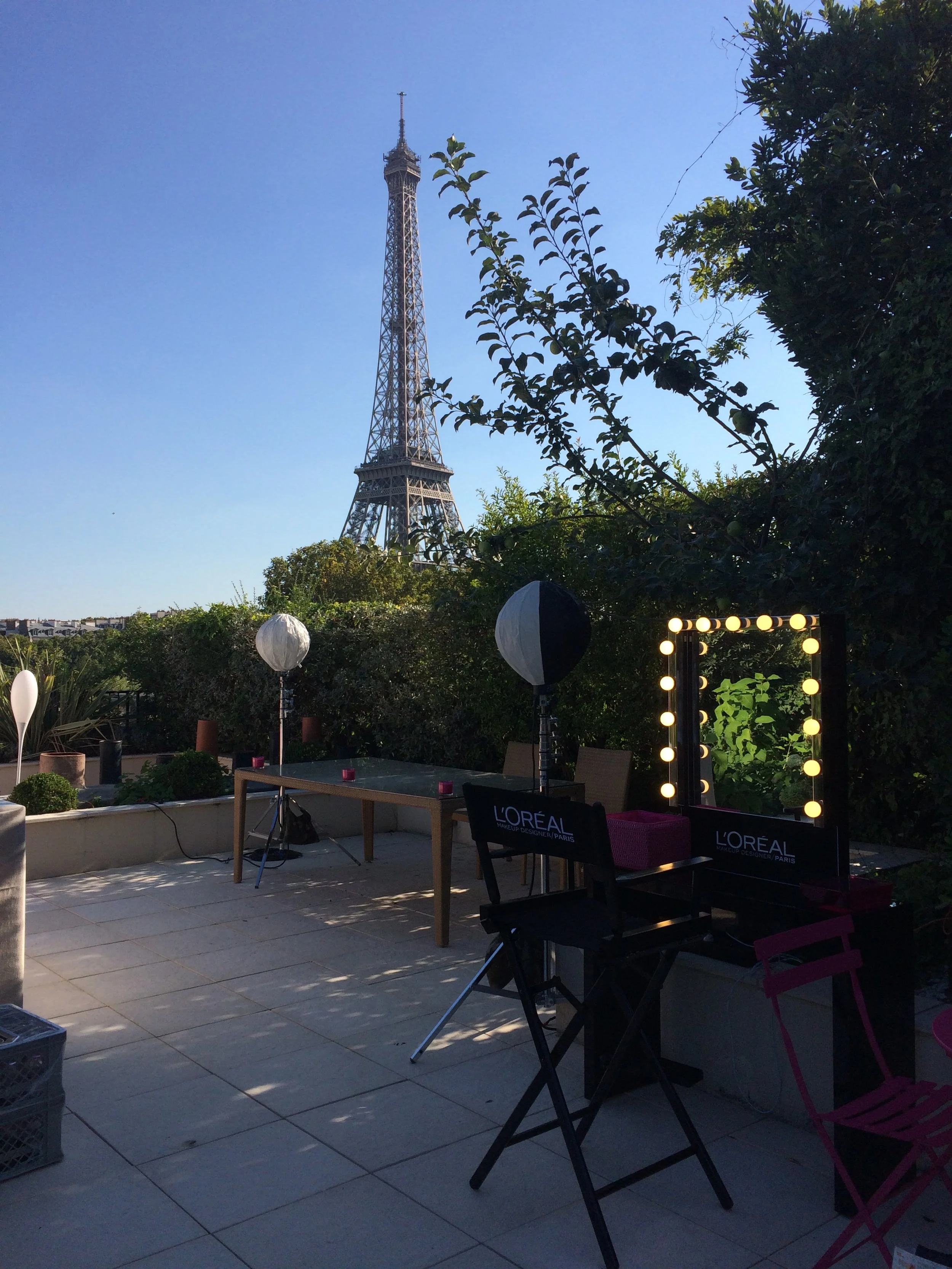 Terrasse avec un décor de maquillage, un miroir éclairé, des lampions, et la Tour Eiffel en arrière-plan dans un ciel clair.