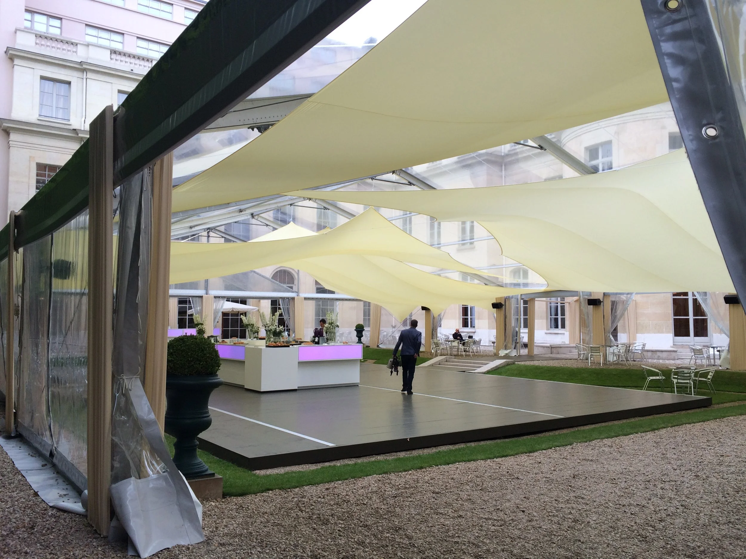 Un espace extérieur décoré pour un événement, avec un toit en tissu jaune suspendu, un sol en bois, des tables et chaises en métal, et des plantes en pot.