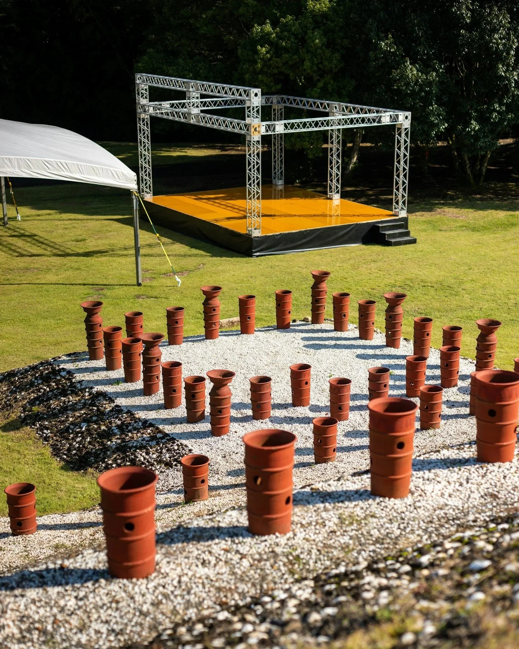 Une scène en plein air avec une scène en métal sur une plateforme orange, entourée d'arbres verts, et plusieurs pots en terre cuite disposés en cercle sur un sol de gravier blanc.