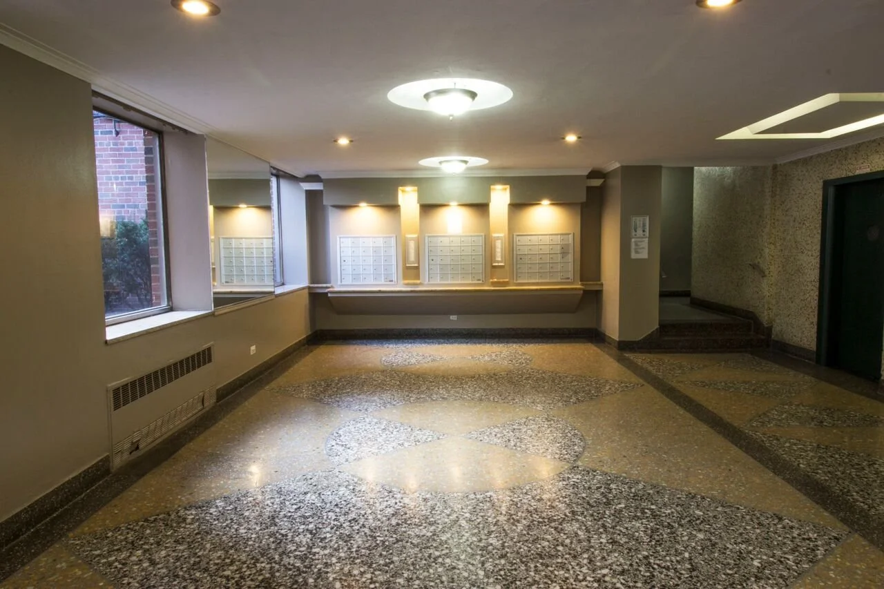 Empty apartment building lobby with mailboxes on the wall, granite flooring pattern, windows to the outside, and an elevator to the right.