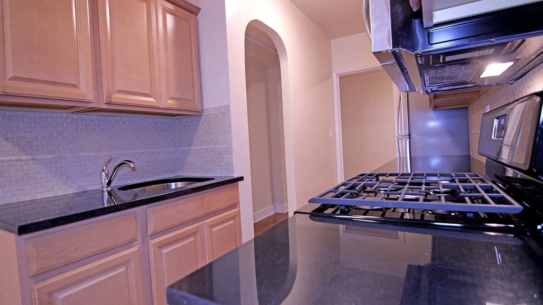 A modern kitchen with light wooden cabinets, a black countertop, a stainless steel stove with a built-in oven, a microwave above the stove, and a small sink with a chrome faucet. There is an entryway visible through an arched opening.