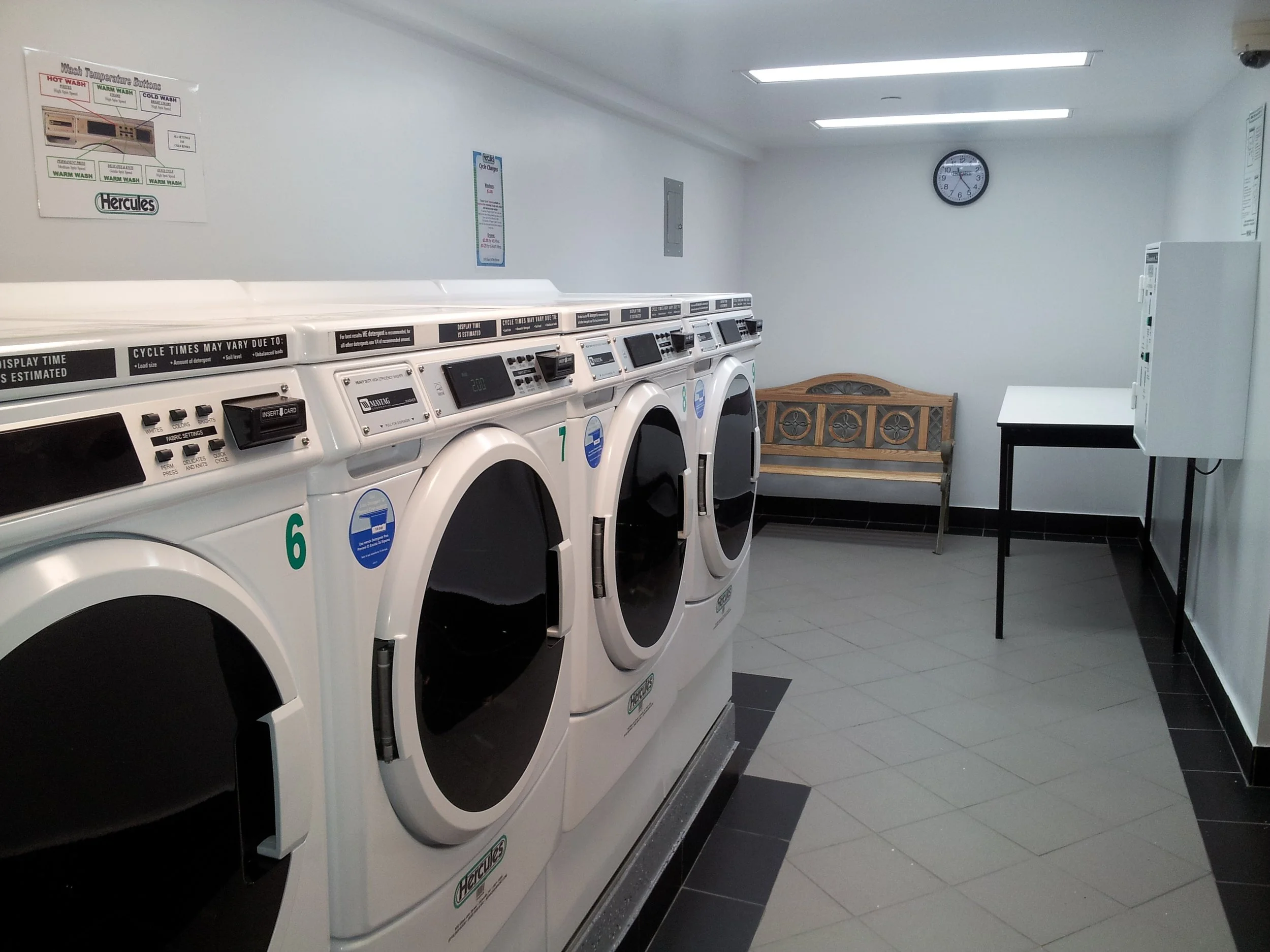 A laundromat with four front-loading washing machines, a wooden bench, a table, and a wall clock.