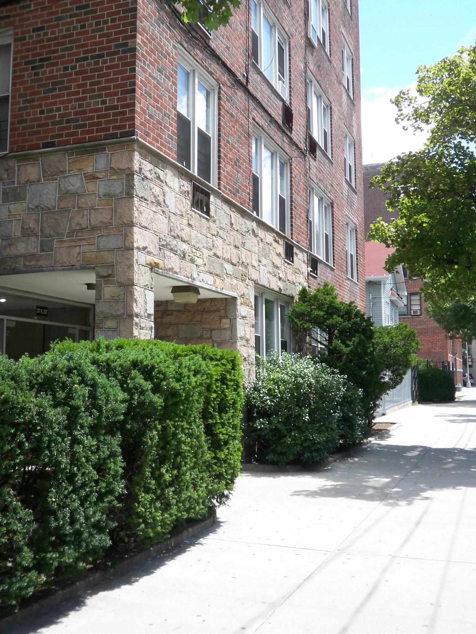 A multi-story brick apartment building with a stone base and surrounding green bushes along the sidewalk.