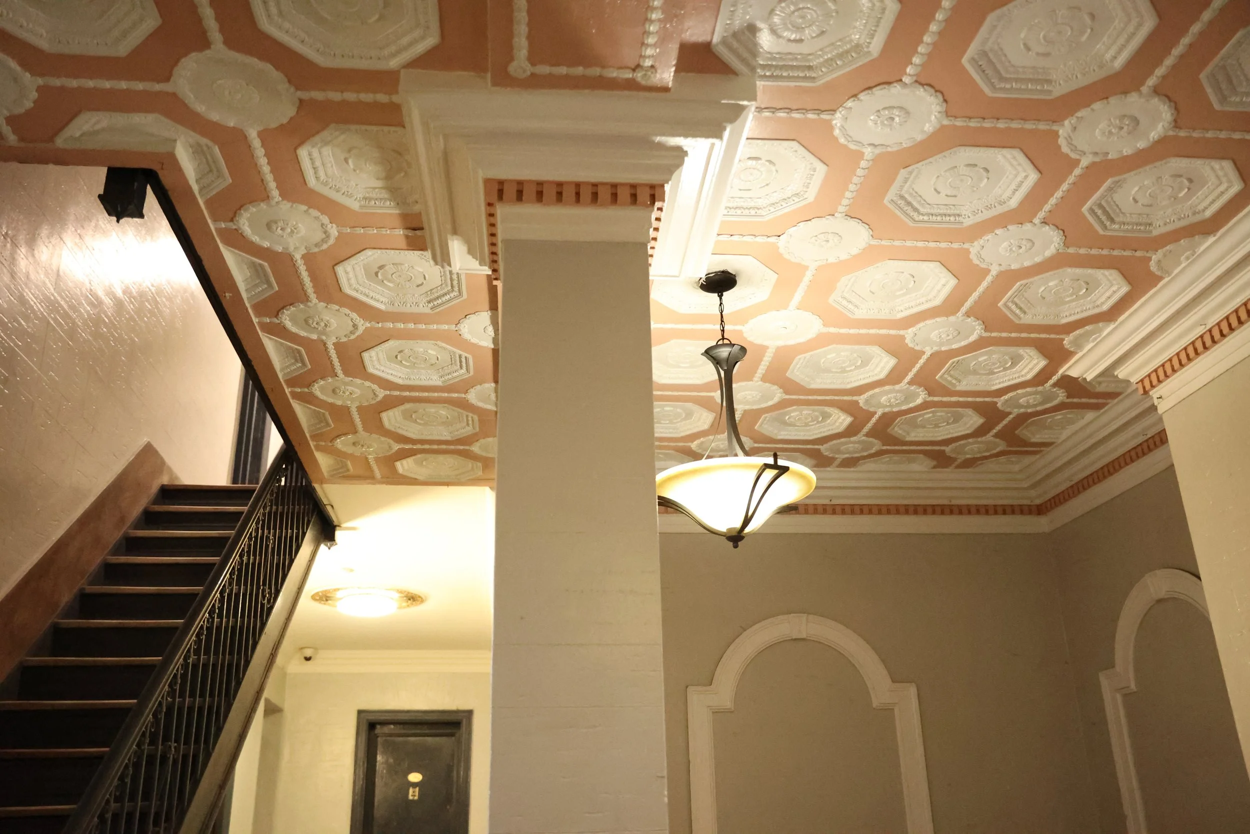 Interior view of a building with a decorative ceiling featuring octagon and floral patterns, a hanging chandelier, a staircase with a dark handrail, and ornate wall moldings.