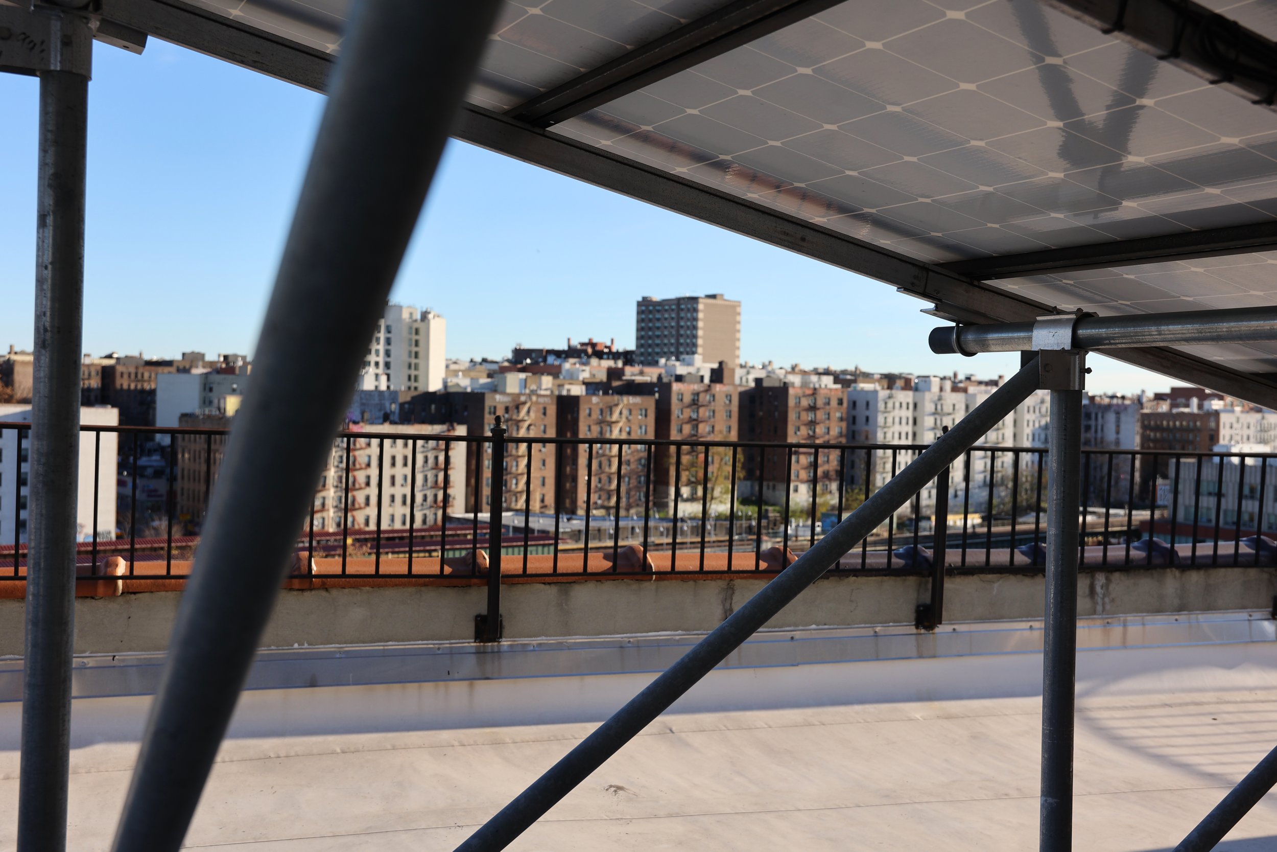 View of a city skyline with multiple apartment buildings seen from a rooftop, partially covered by scaffolding and solar panels.