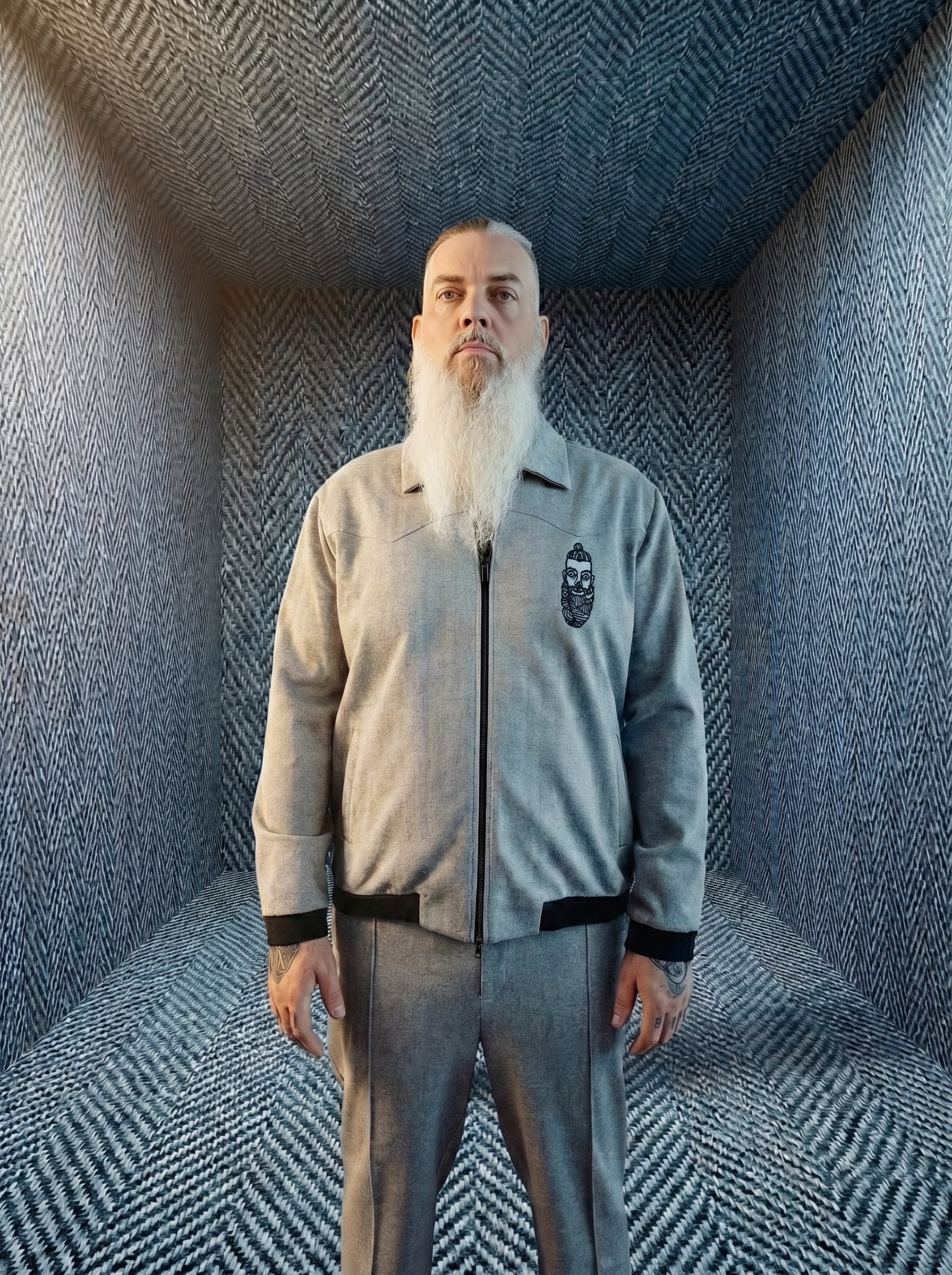 A man with long gray beard and mustache standing in a soundproof room with patterned walls and ceiling, wearing a gray zip-up jacket with a graphic of a bearded man on the chest.