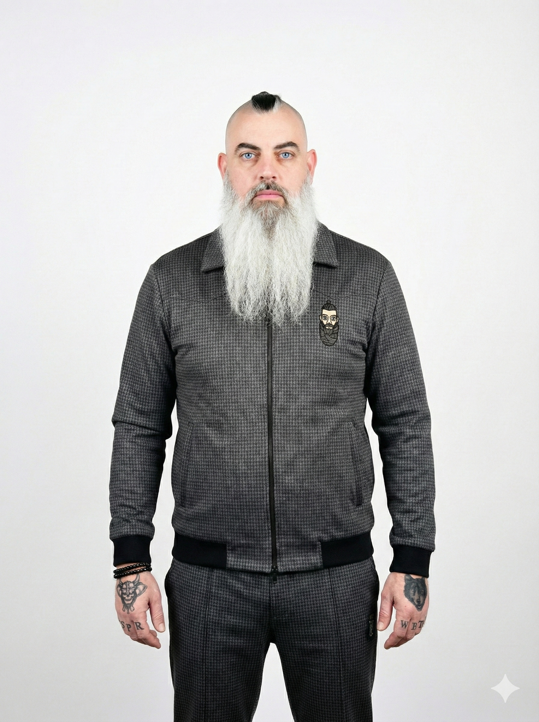 A man with light skin, blue eyes, and a long gray beard, wearing a dark gray checkered zip-up jacket with a small embroidered face logo on the chest. He has tattoos on his hands, including a face tattoo on his right hand and words on his fingers, and wears black bracelets on his left wrist. The background is plain white.