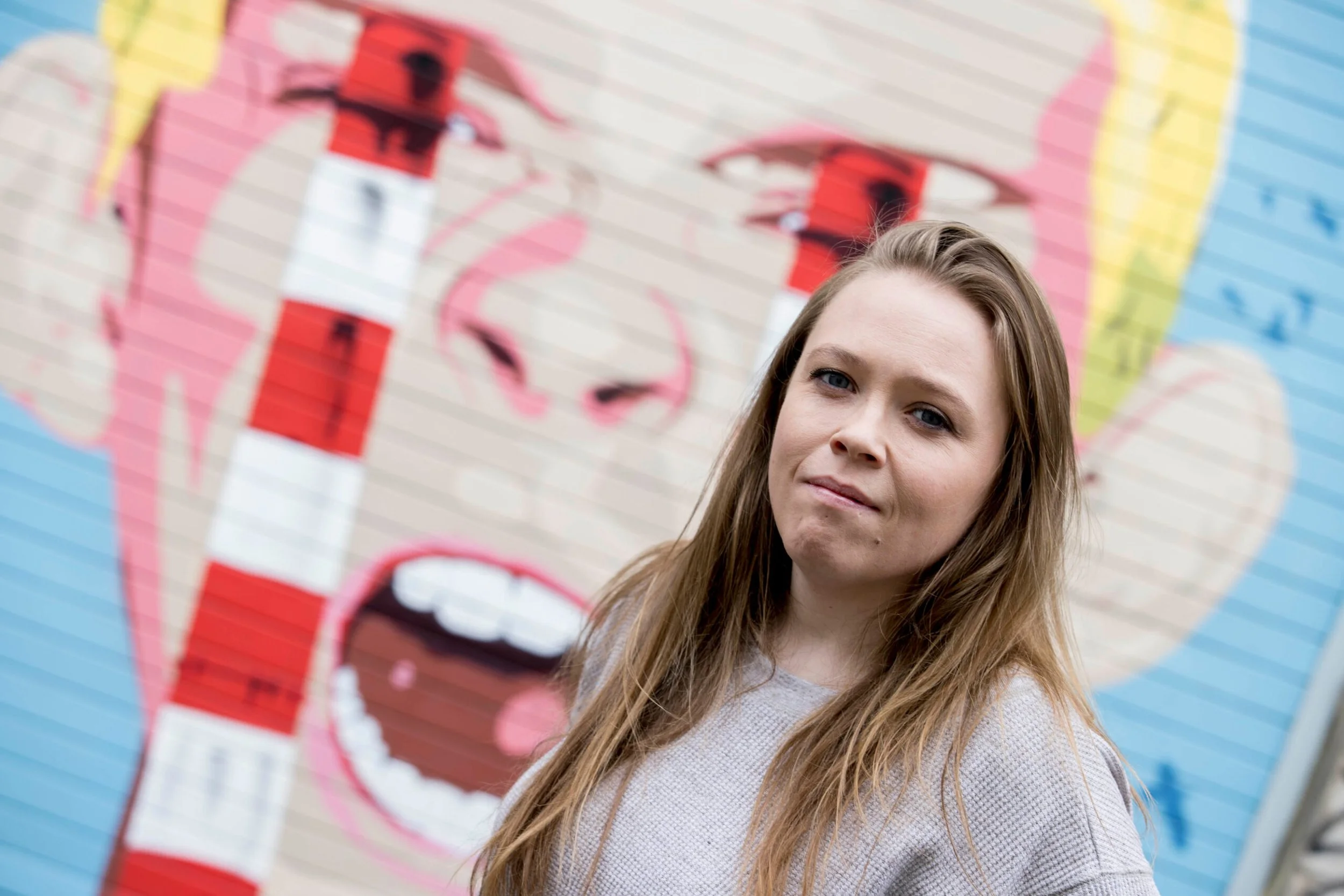 A woman with long brown hair stands in front of colorful graffiti art on a brick wall, featuring a stylized face with pink cheeks and lips, and blue and yellow background elements.