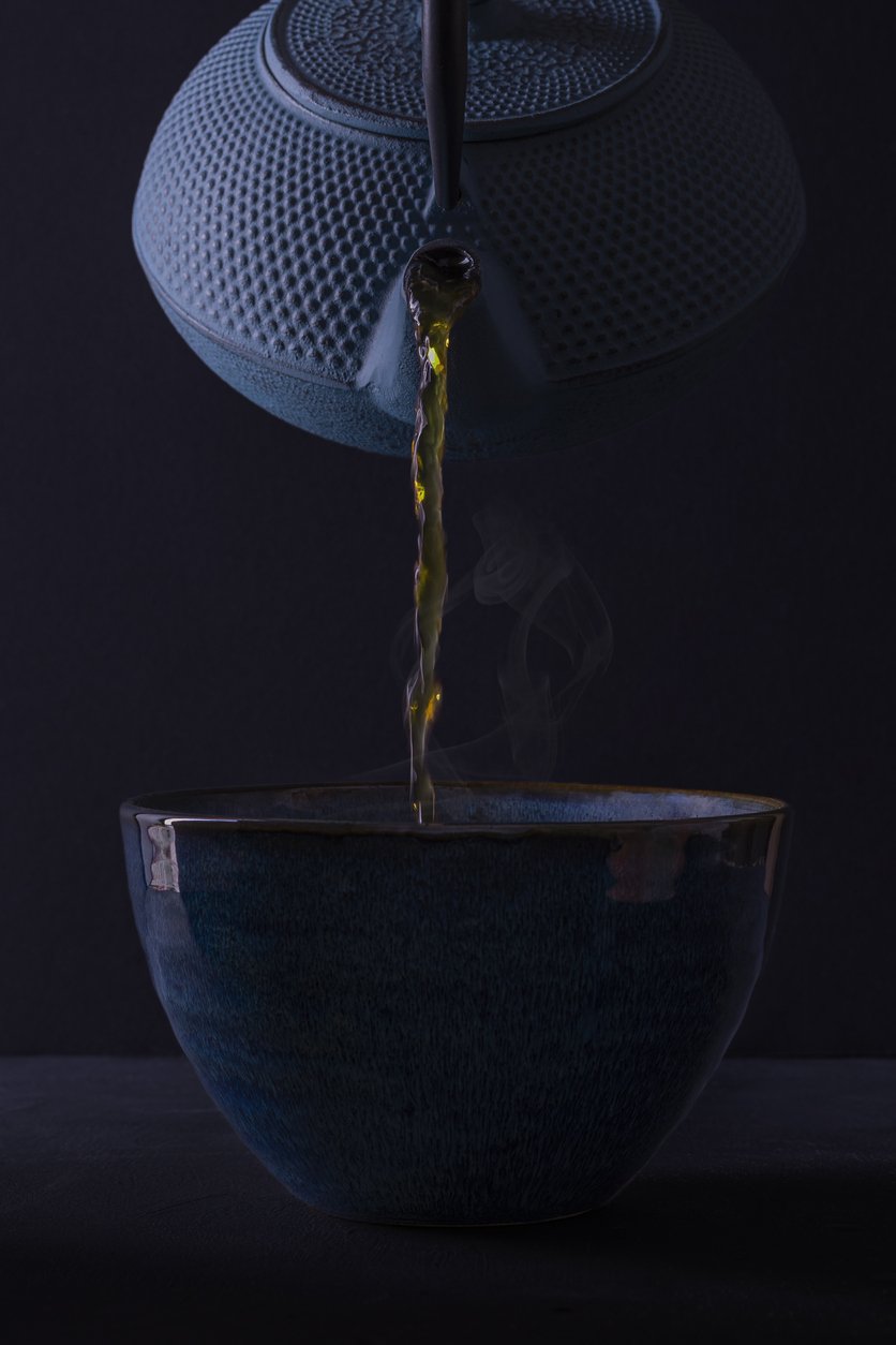 A blue teapot pouring hot tea into a dark blue bowl on a black background.