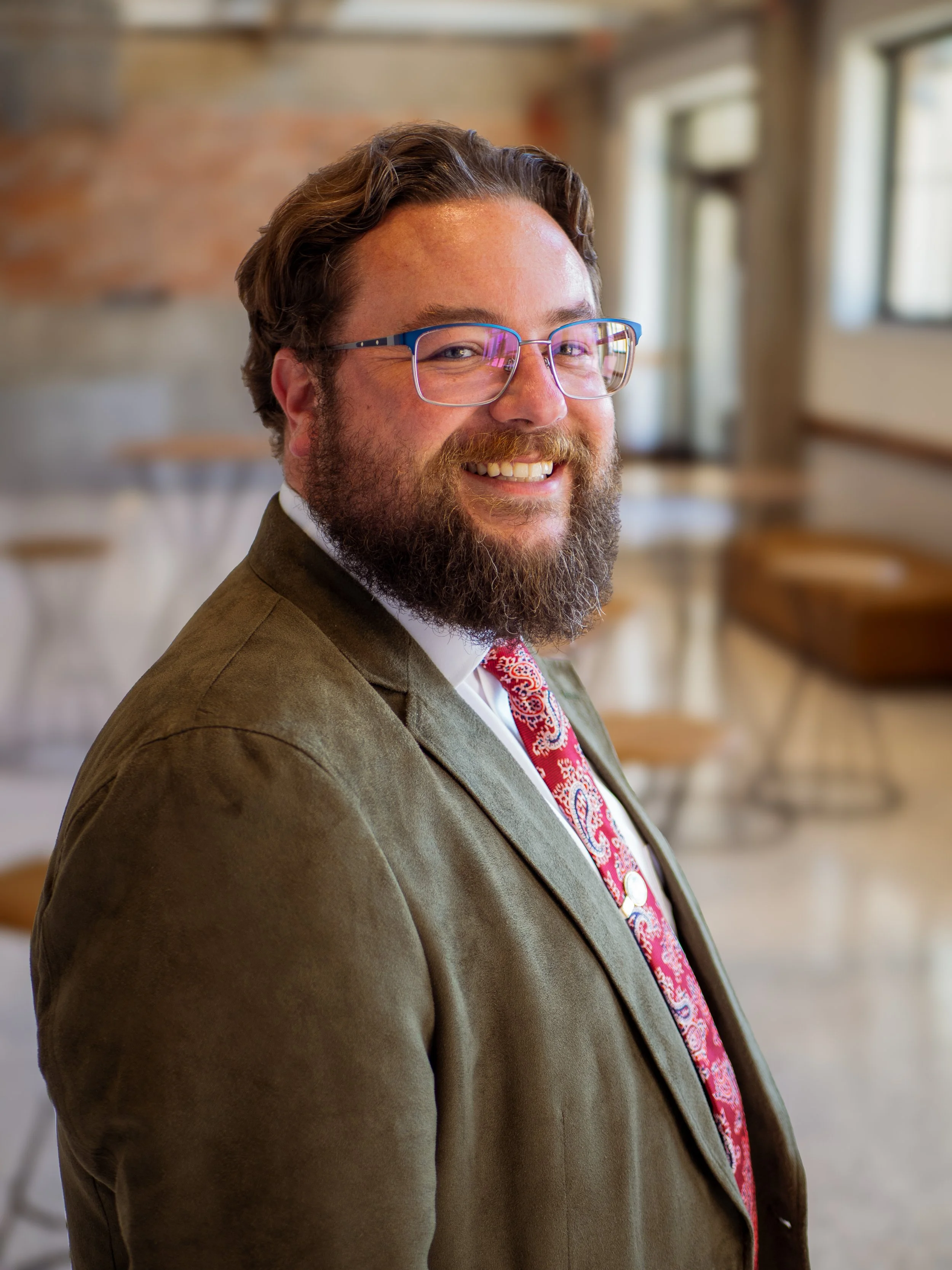 A smiling bearded man wearing glasses, a suit, and a patterned tie, standing in a well-lit room with large windows and wooden furniture.