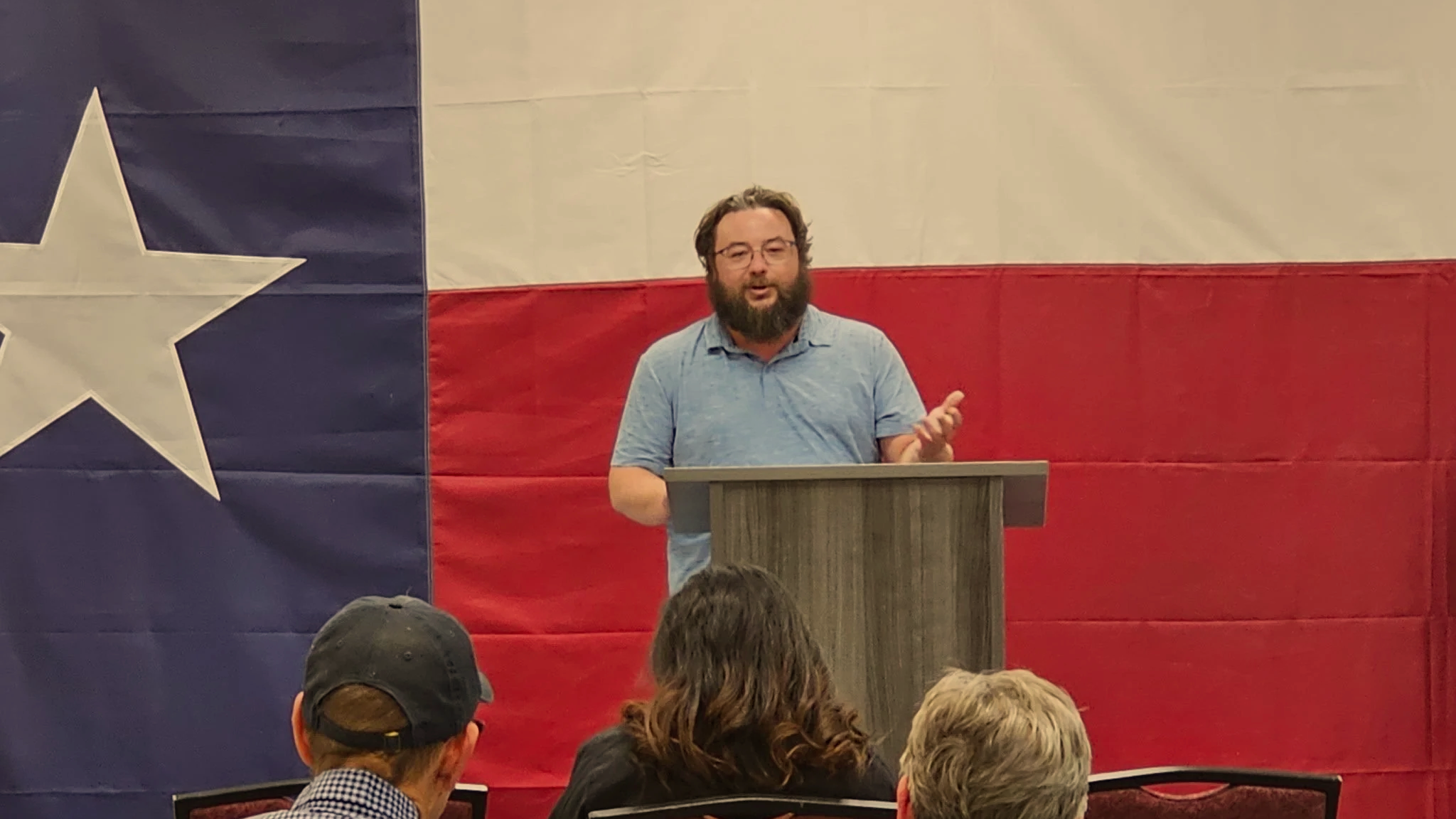 A man with glasses and a beard is standing behind a wooden podium, speaking to an audience, in front of a large Texas flag.
