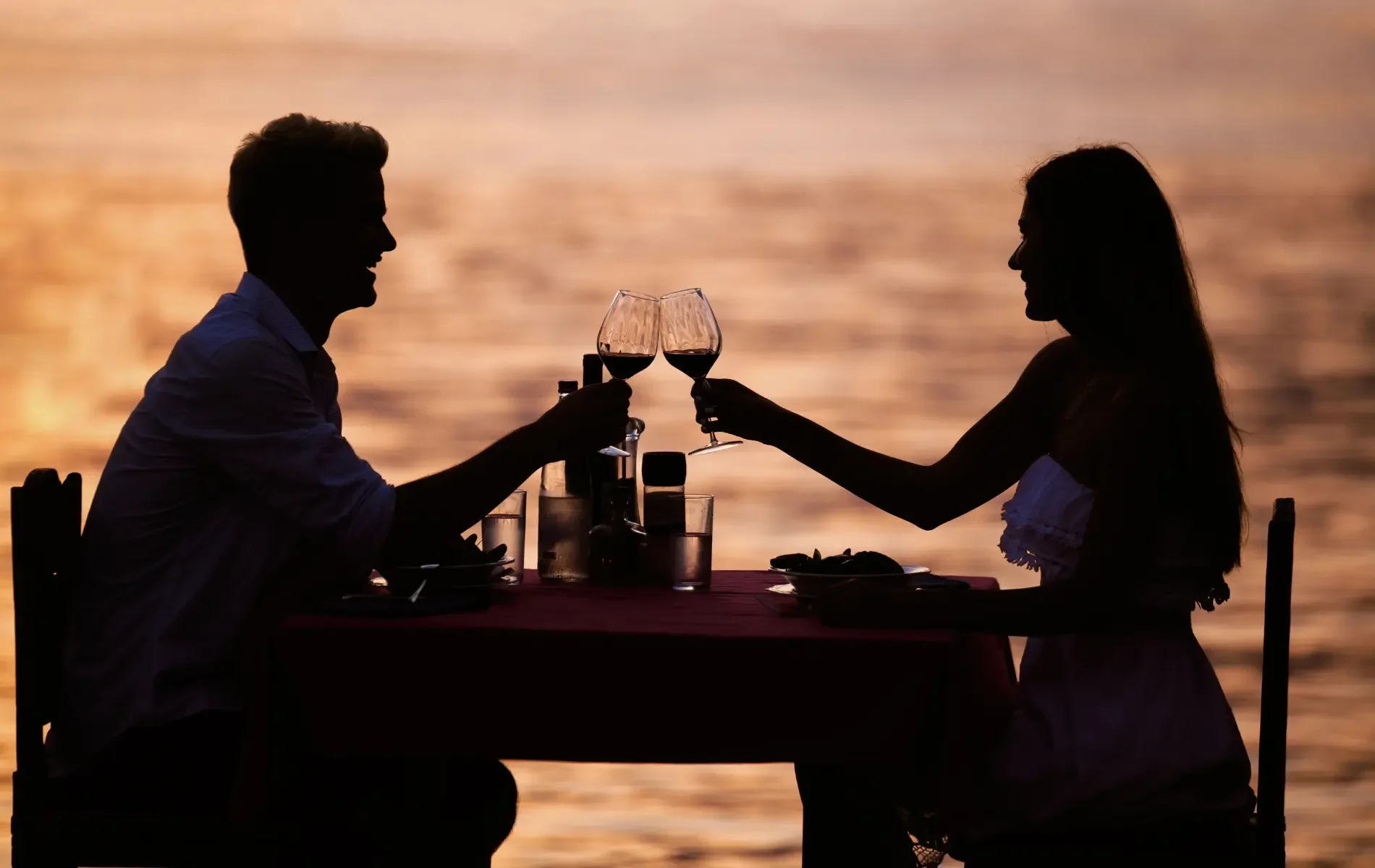 Silhouette of a man and woman clinking wine glasses at an outdoor dinner table by the water during sunset.
