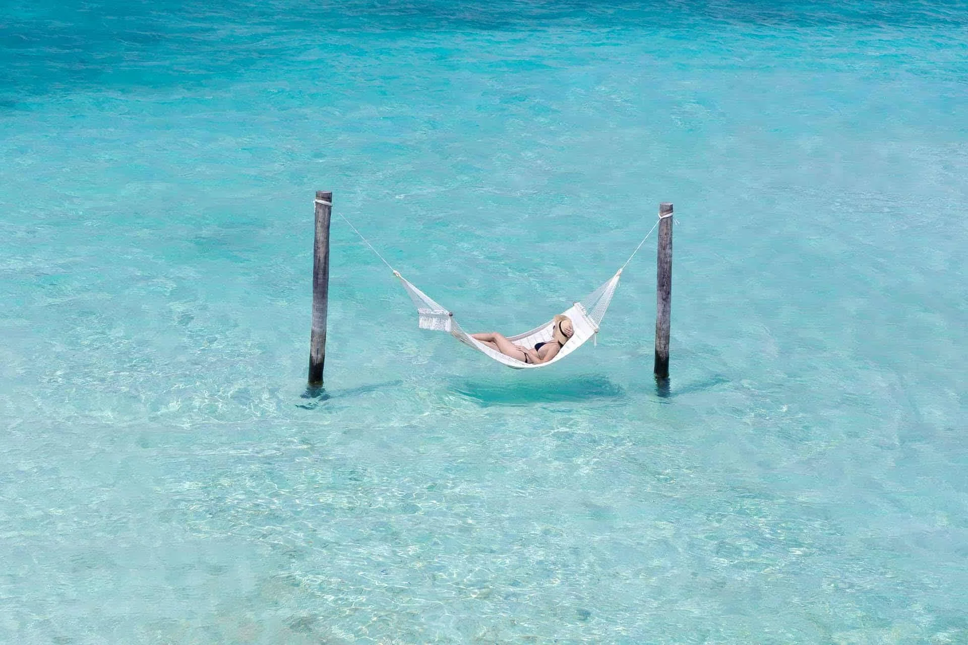 A woman relaxing in a hammock strung between two wooden posts in clear turquoise water.
